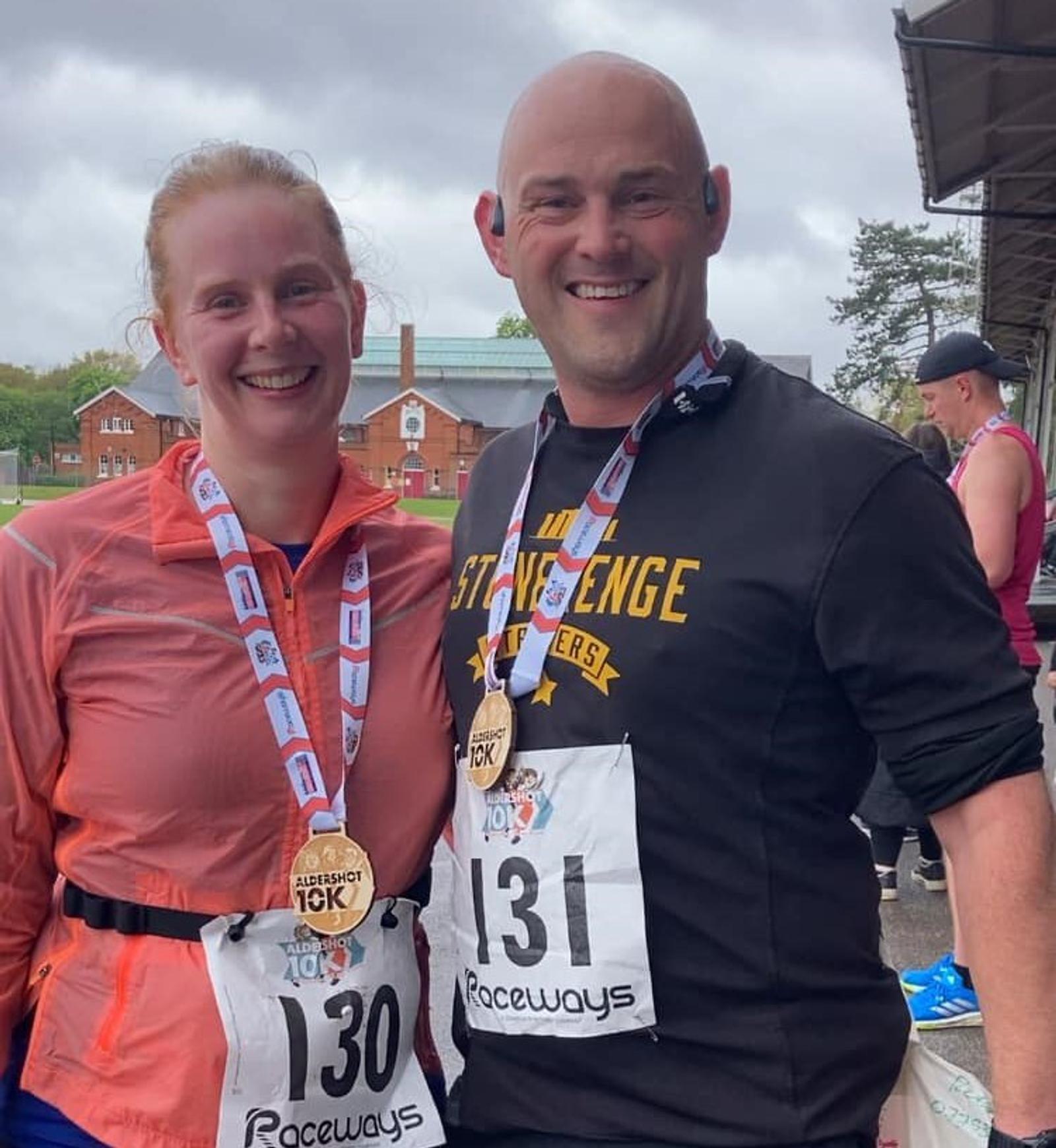 Two smiling runners wearing numbered race bibs and medals around their necks pose together. The woman wears a pink jacket and the man a black shirt. They stand under an overcast sky with buildings and other runners in the background.