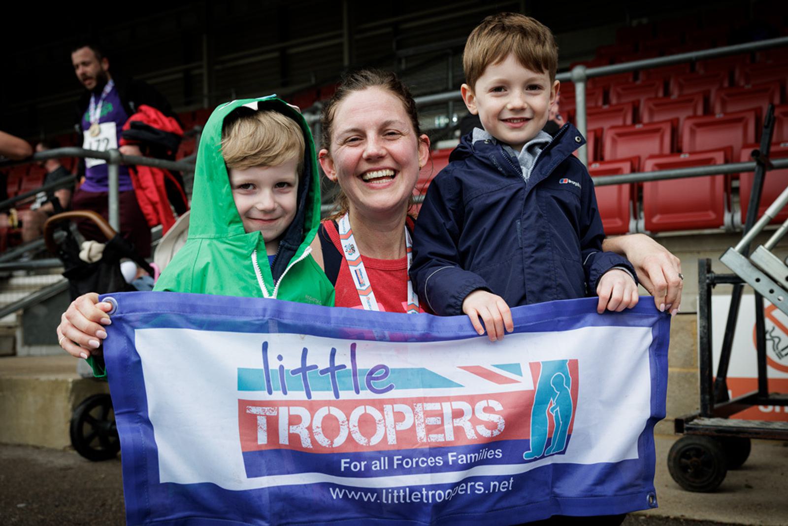 A smiling woman with a medal around her neck is holding a "Little Troopers" banner alongside two children. They are standing in a stadium with red seats in the background. One child is wearing a green jacket, and the other is in a blue jacket.