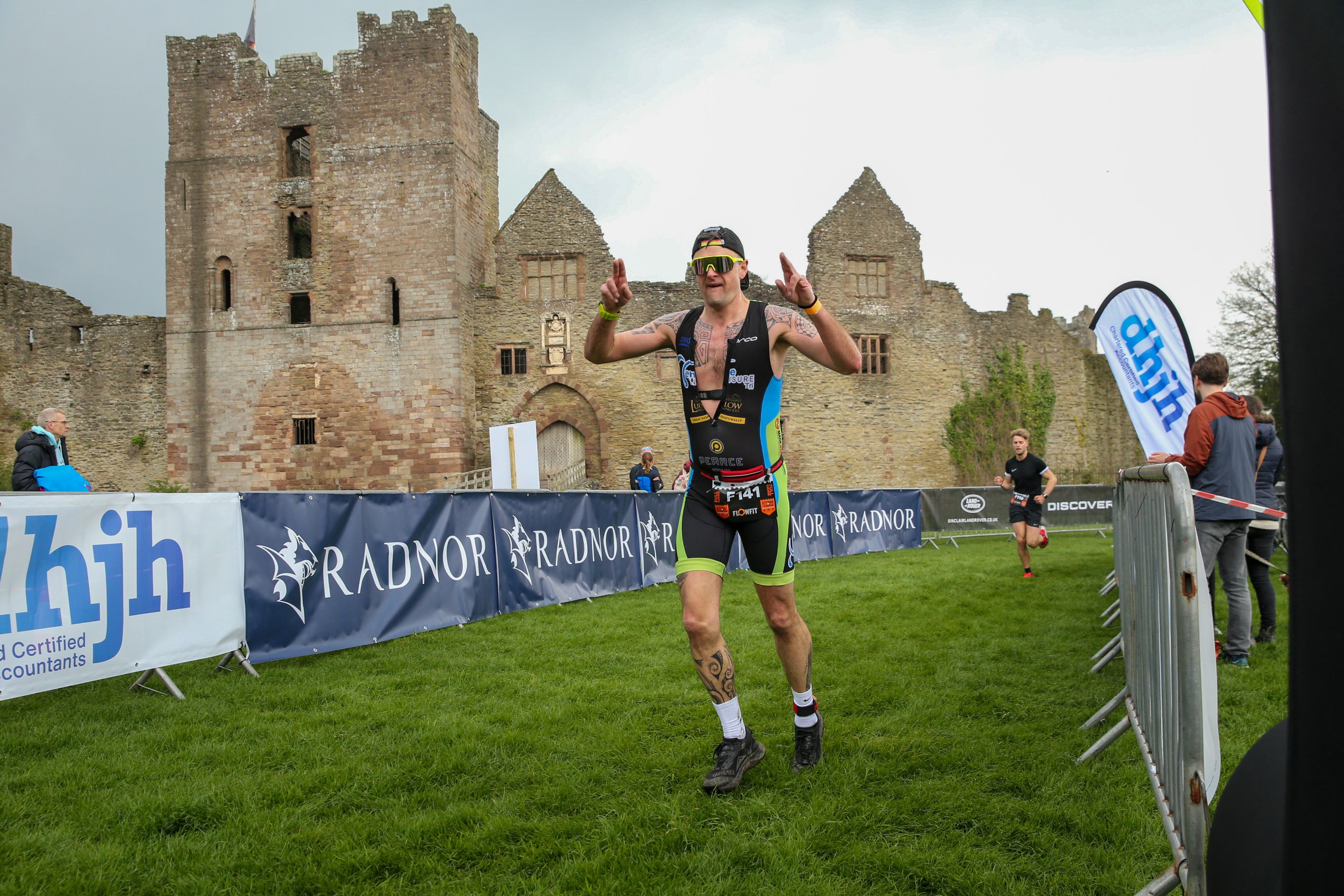 A triathlete wearing a black and green outfit runs past a historic stone building, raising his arms in celebration. Banners line the grassy path, and a few onlookers stand nearby.