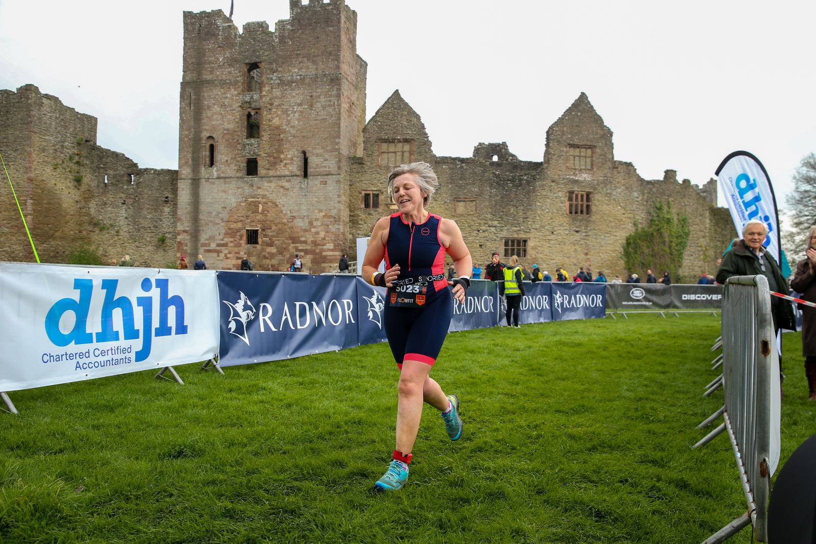 A woman wearing a triathlon suit runs past a historic castle and banners during a race. The grass is green, and the sky is overcast. Spectators watch nearby.