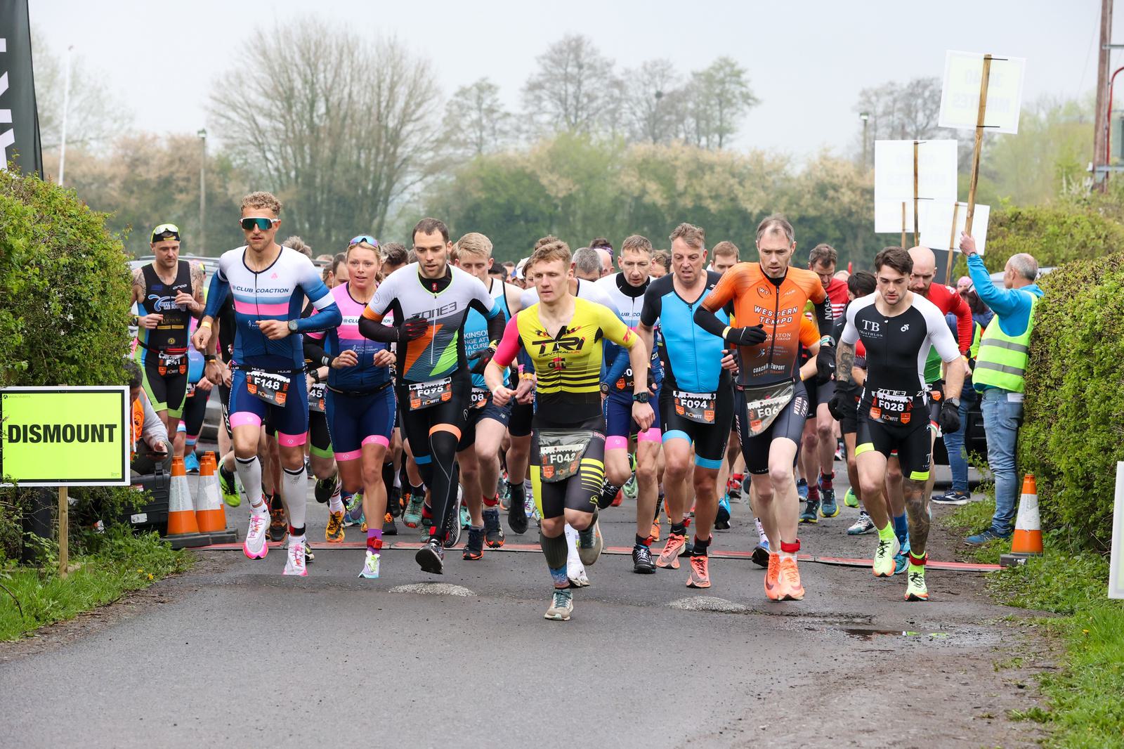 A group of athletes in vibrant sports gear are running on a road during a race. They are surrounded by greenery and road signs, with some spectators in the background. The energy is high as they start the competition.