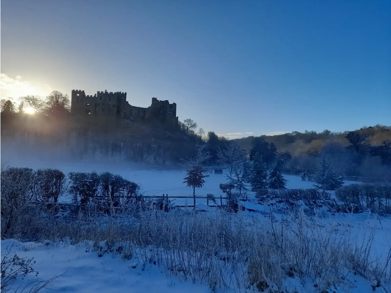 A snow-covered landscape with a large, ancient castle in the background. The sun is setting behind the trees, casting a warm glow over the scene. Bushes and trees are lightly dusted with snow in the foreground.