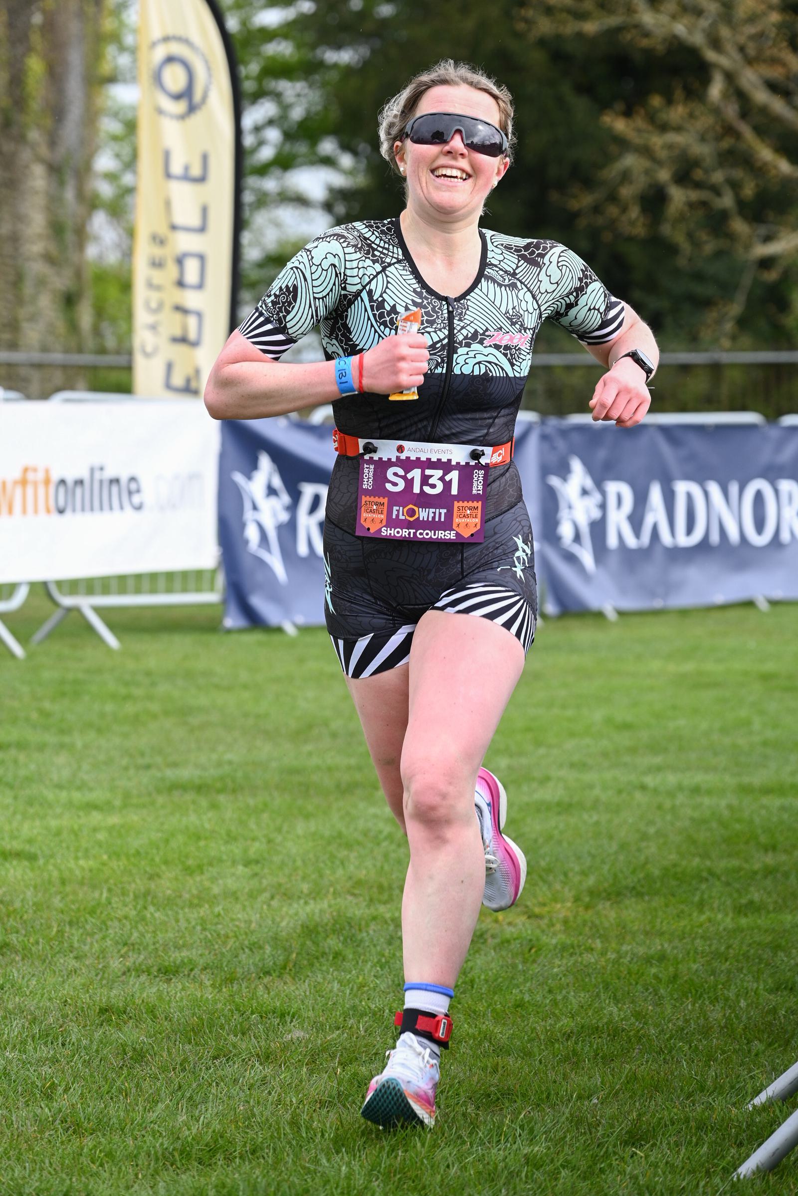 A person is running outdoors on grass, wearing athletic gear and a race bib labeled "S131." They are smiling, with sunglasses and short hair. There are banners and barriers in the background.