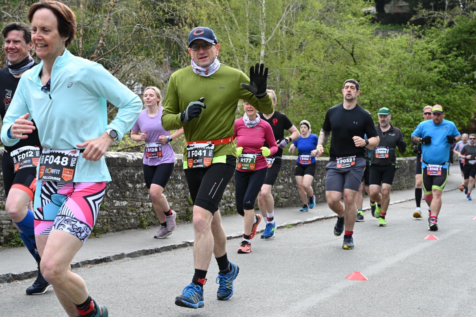 A group of runners in colorful athletic gear race along a street lined with trees. They appear determined and focused. The race numbers are visible on their shirts, and some participants wear caps and sunglasses.