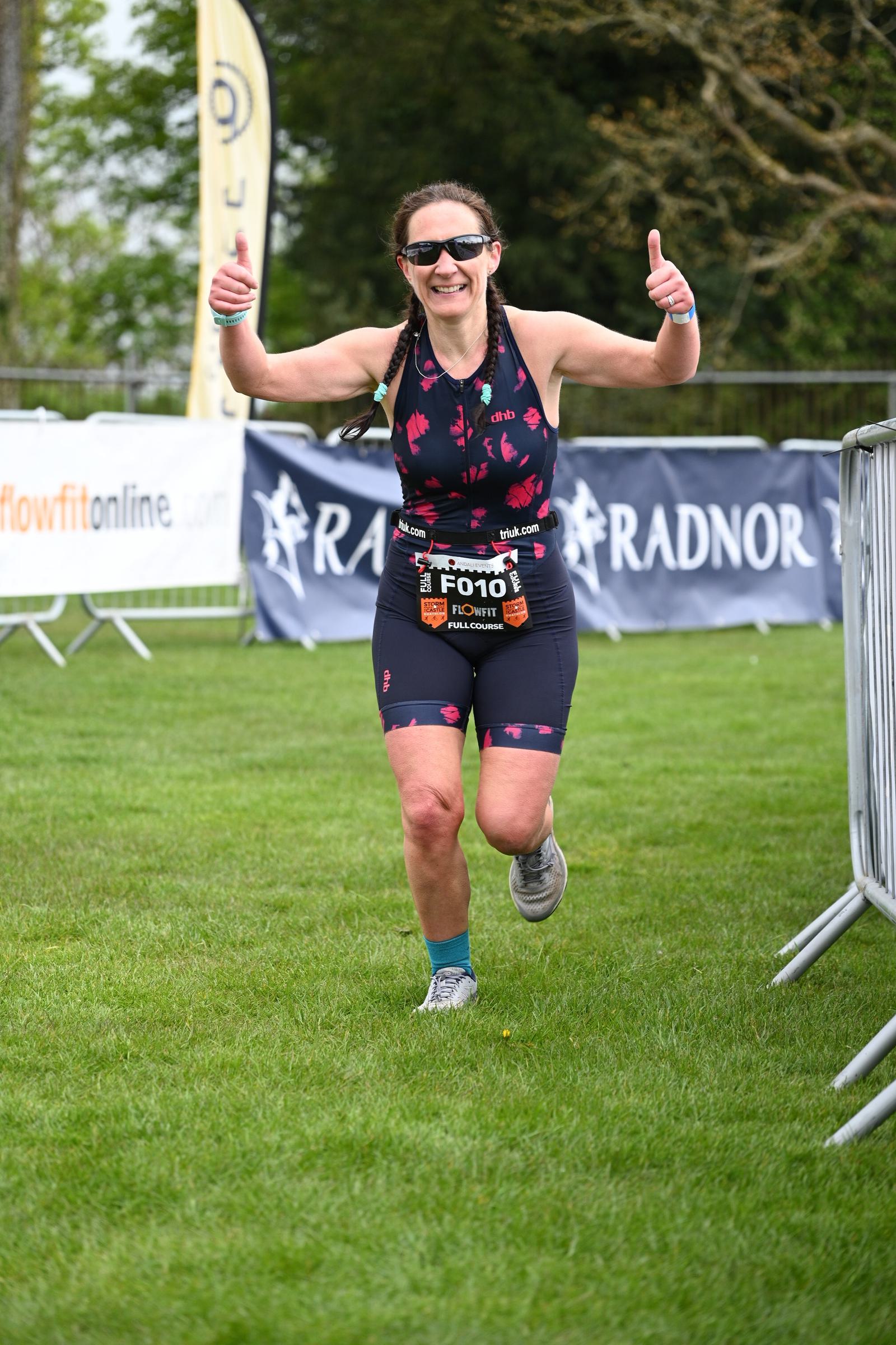 A person wearing athletic gear is running on grass, smiling and giving thumbs up. They have a race bib and sunglasses. A banner and metal barriers are in the background.