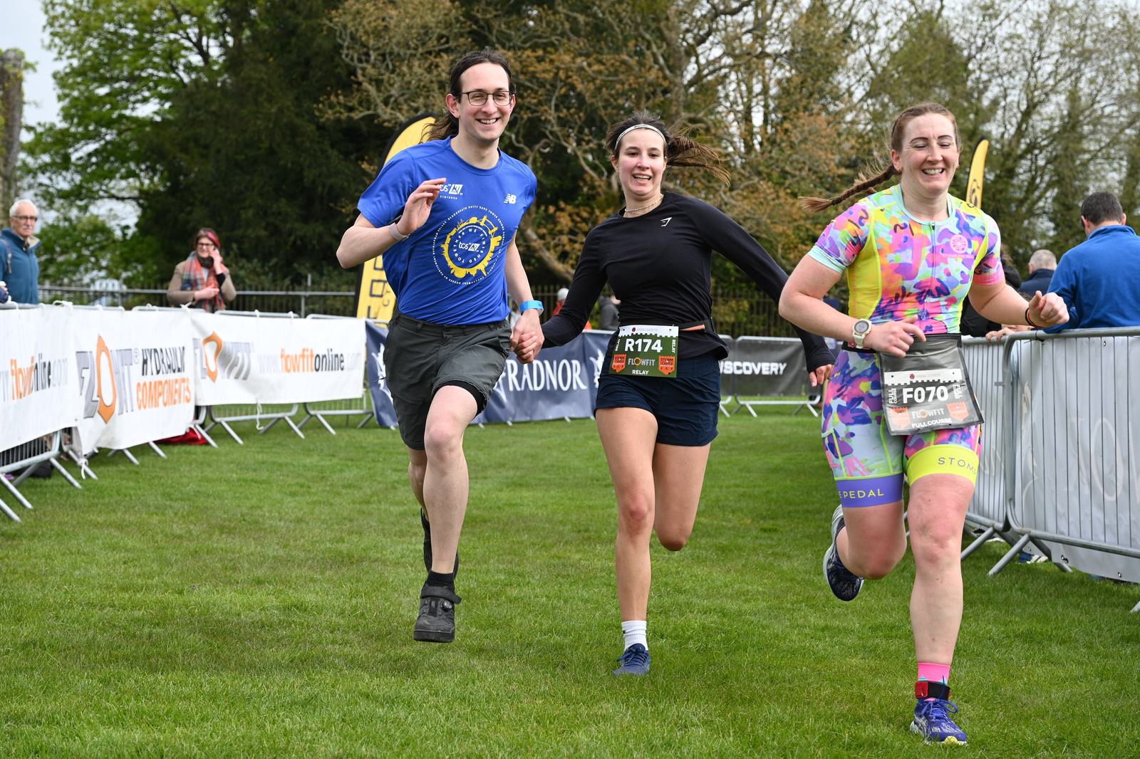 Three people are running and smiling as they approach the finish line of a race. They are on grass with race banners in the background. Spectators are visible behind the barriers.