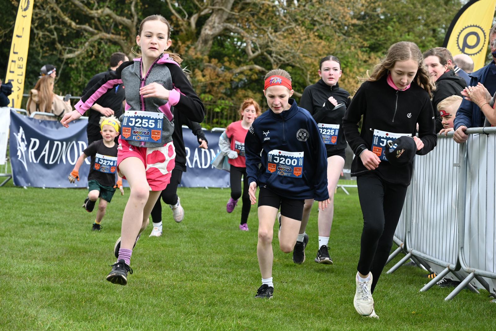 A group of children running on grass during a race event, with some wearing race bibs and sports attire. Spectators are beside the course, behind metal barriers. Trees are visible in the background.