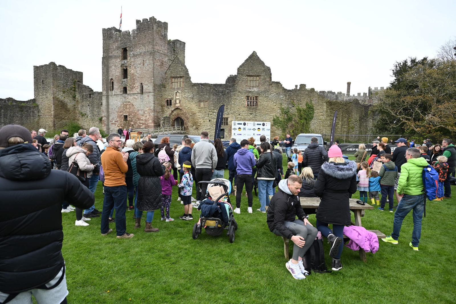 A crowd of people gathers on a grassy field in front of a large, ancient stone castle. Some people sit at a wooden picnic table, while others stand and converse. There are banners and cars in the background. The sky is overcast.