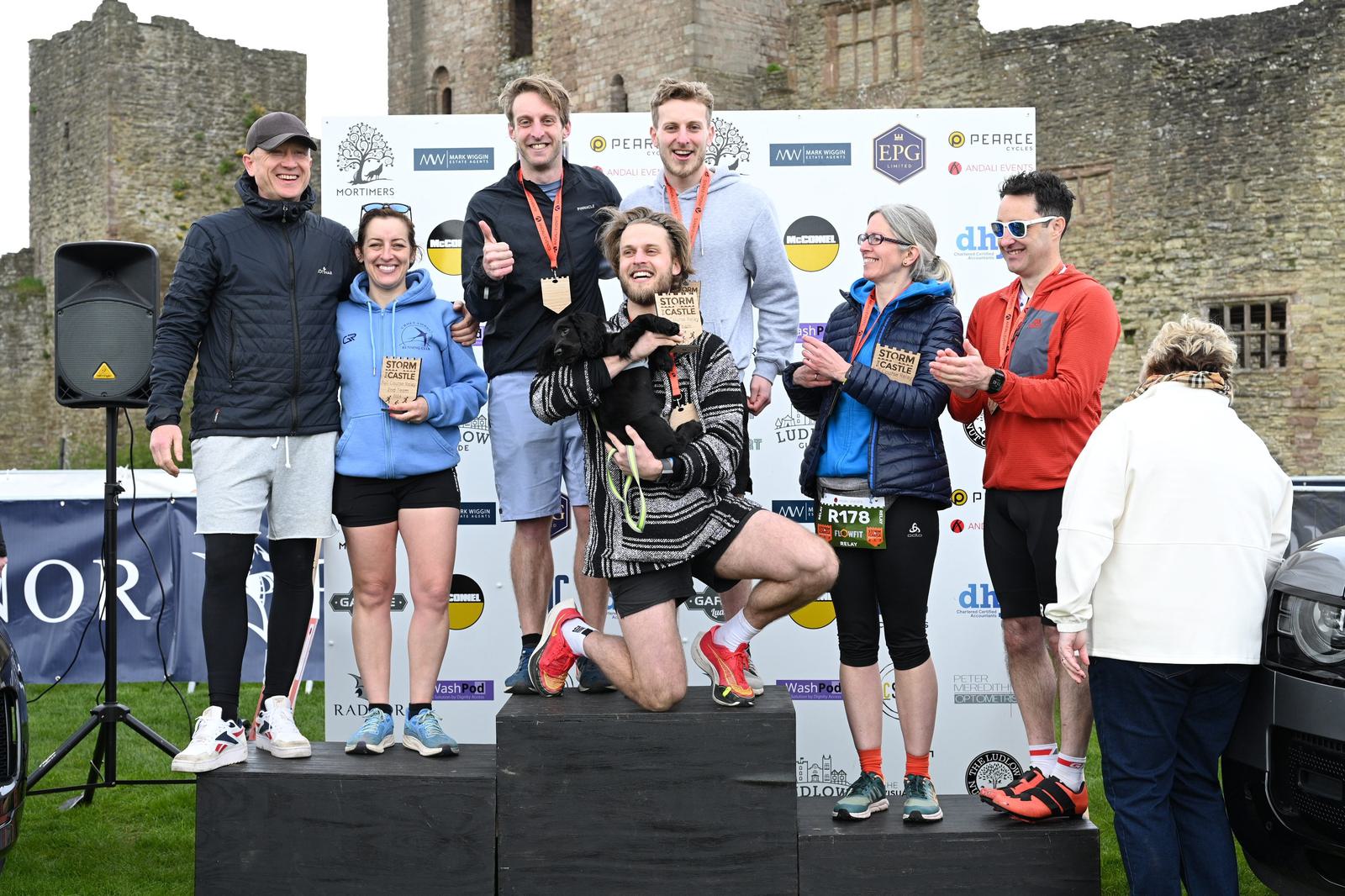 Seven people are celebrating on a podium in front of a stone building, some wearing race medals and holding awards. They are dressed in athletic wear, and there is a small dog being held by one person. A large banner is in the background.
