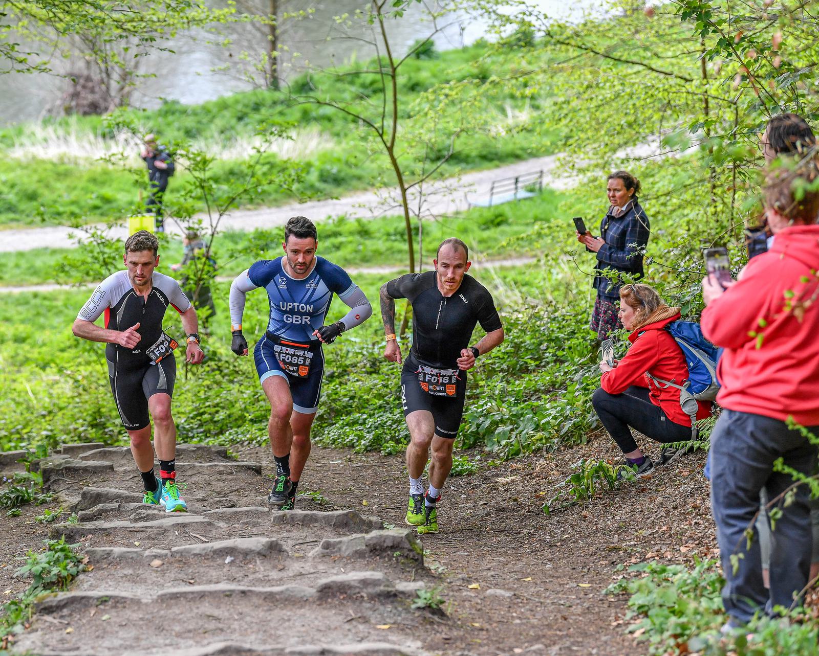 Three athletes wearing race bibs run uphill through a wooded area on a trail. Two men on the left wear black shorts and shirts, while the man in the middle wears a blue suit. Spectators watch and take photos along the path.
