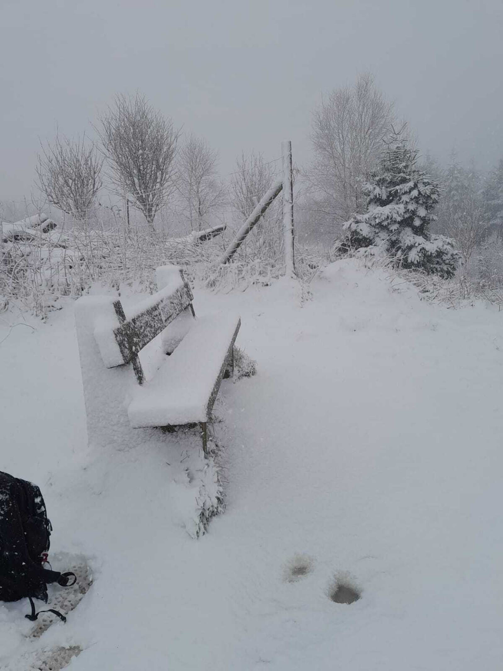 A snowy scene with a bench covered in snow, surrounded by snow-covered trees and a fence. Footprints are visible on the ground, and a backpack rests nearby. The sky is overcast, adding to the wintery atmosphere.