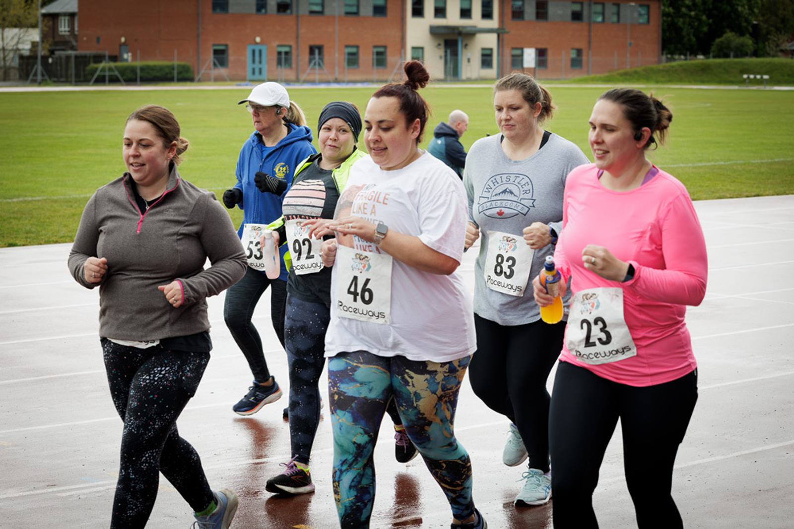 A group of women jogging together on a wet track, wearing race numbers and athletic gear. The background shows a grassy field and a building. They appear focused and are at different paces.