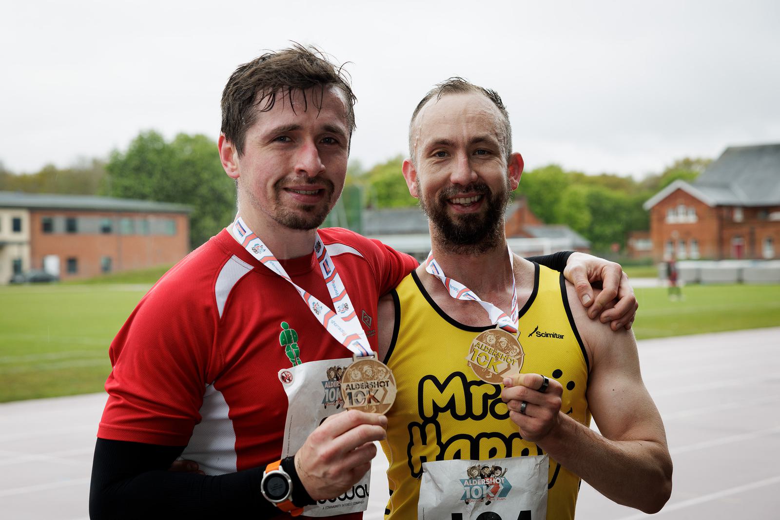 Two men smiling and holding medals pose for a photo on a track. One wears a red shirt and the other a yellow shirt. Both have race bibs on their shirts and medals around their necks, celebrating their achievement.