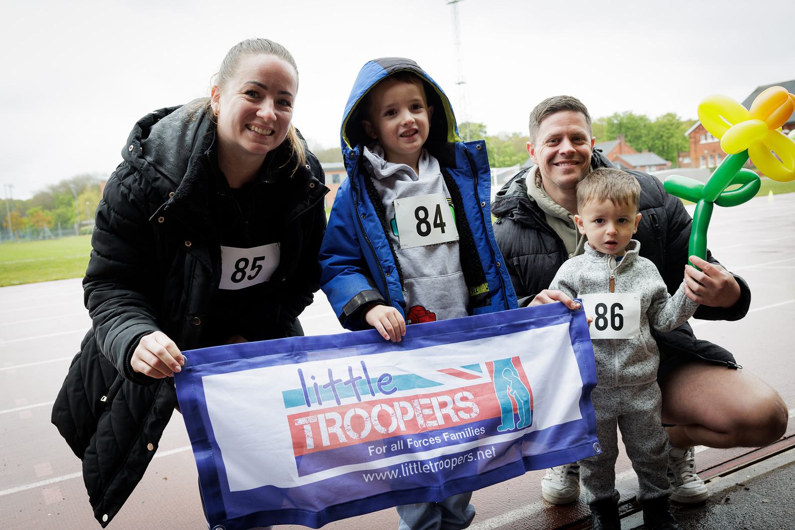 A family of four stands together on a track. They hold a "Little Troopers" banner. The woman and children wear numbered bibs. One child holds a yellow balloon shaped like a flower. Everyone is dressed in warm clothing.