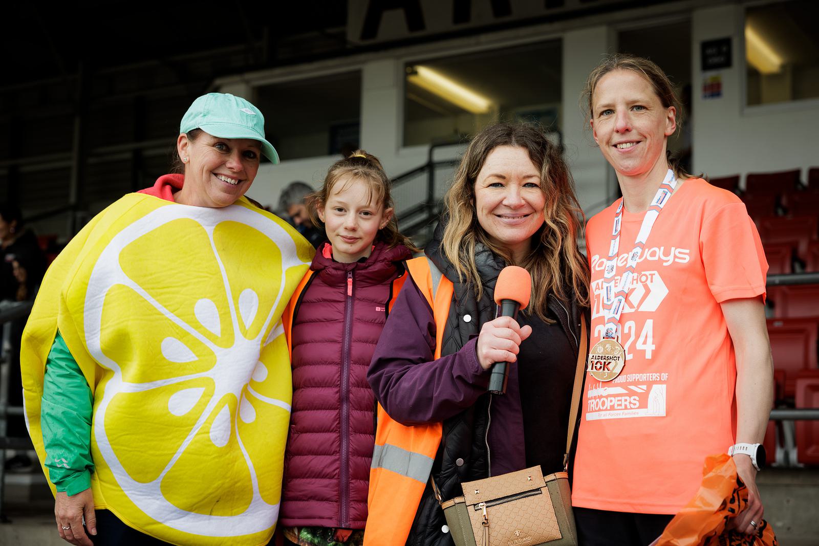 Four people are posing for a photo. One person is dressed in a lemon costume, another in an orange T-shirt with a medal. A third person is holding a microphone and wearing an orange vest, and a young girl is in casual wear. They are in a stadium.
