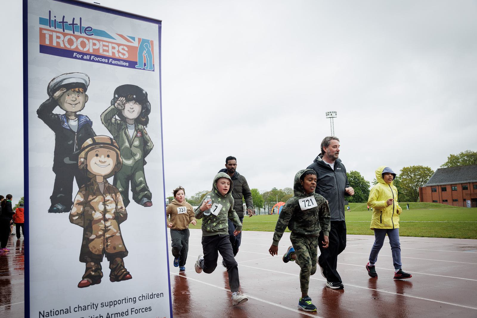 Children and adults run on a wet track in a race. A sign on the left displays "Little Troopers" with cartoon soldiers. The sky is overcast, and a grassy field and building are in the background.