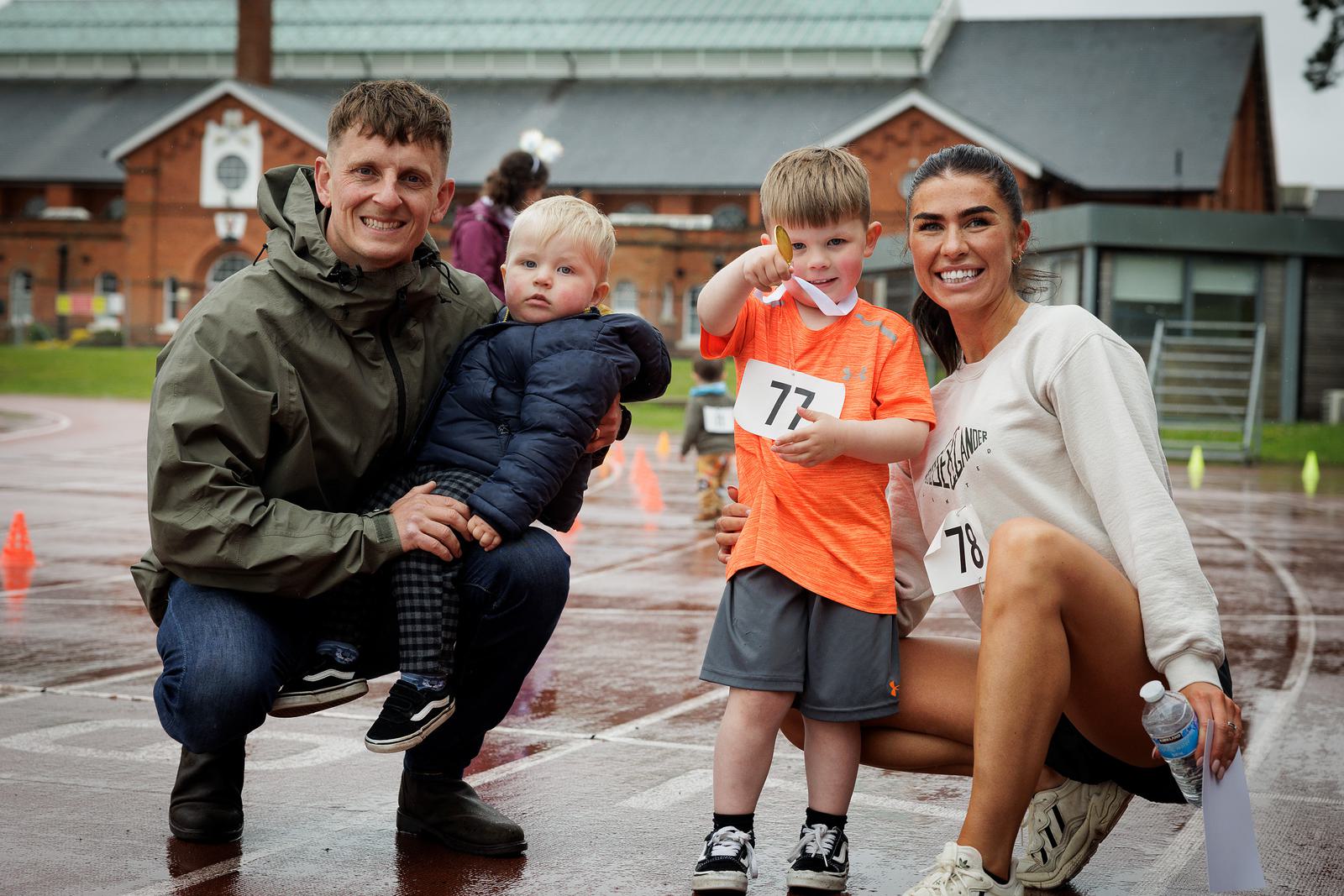 A man and woman crouch on a wet track with two young children, one in each of their arms. The woman holds a water bottle, and the boy next to her points forward. Both children wear numbered bibs. The background shows a track field and a building.