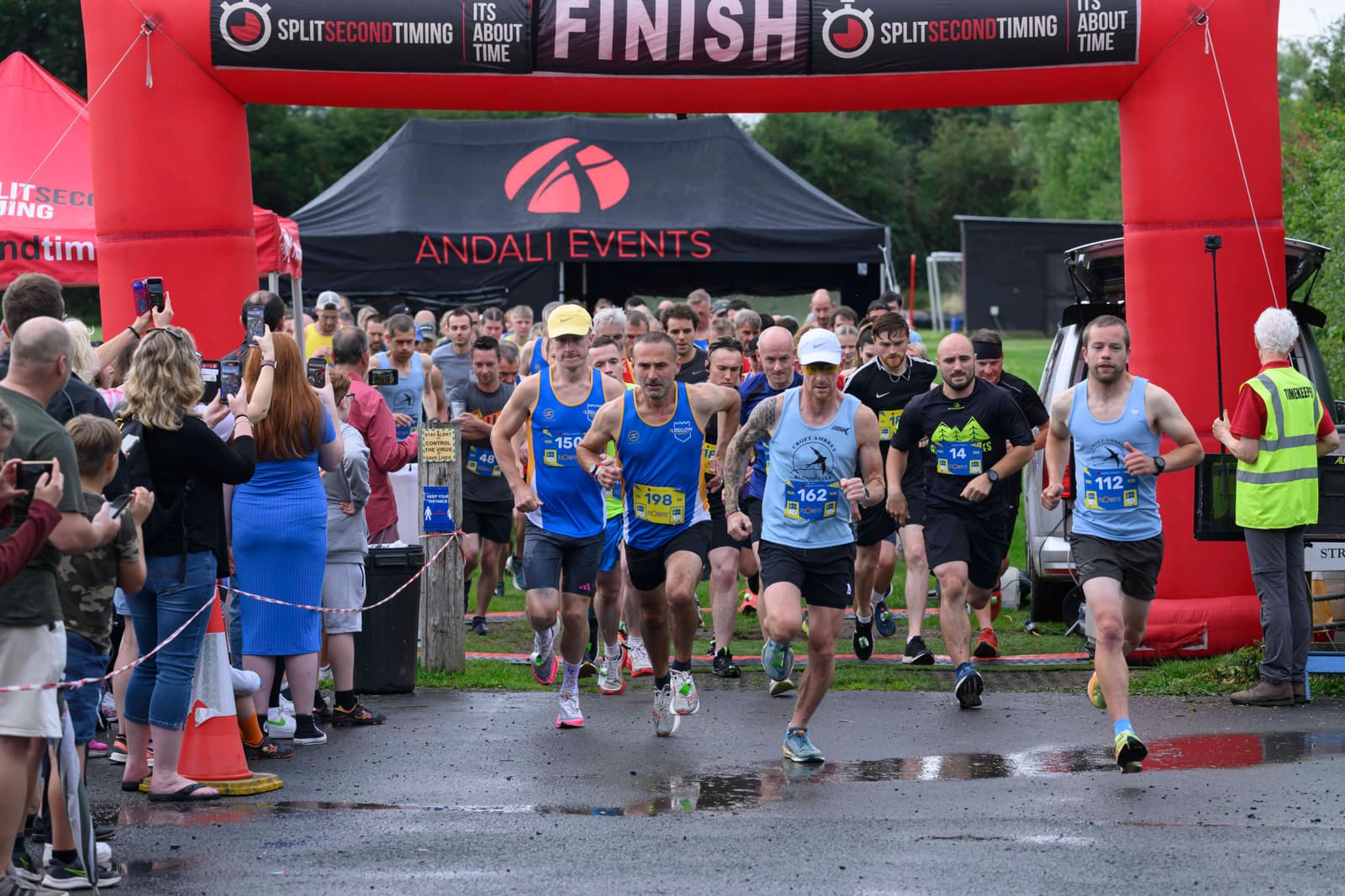 Runners in blue shirts and race bibs begin a race at a starting line under a red arch, while a crowd of spectators watches and takes photos. Trees and tents are visible in the background.
