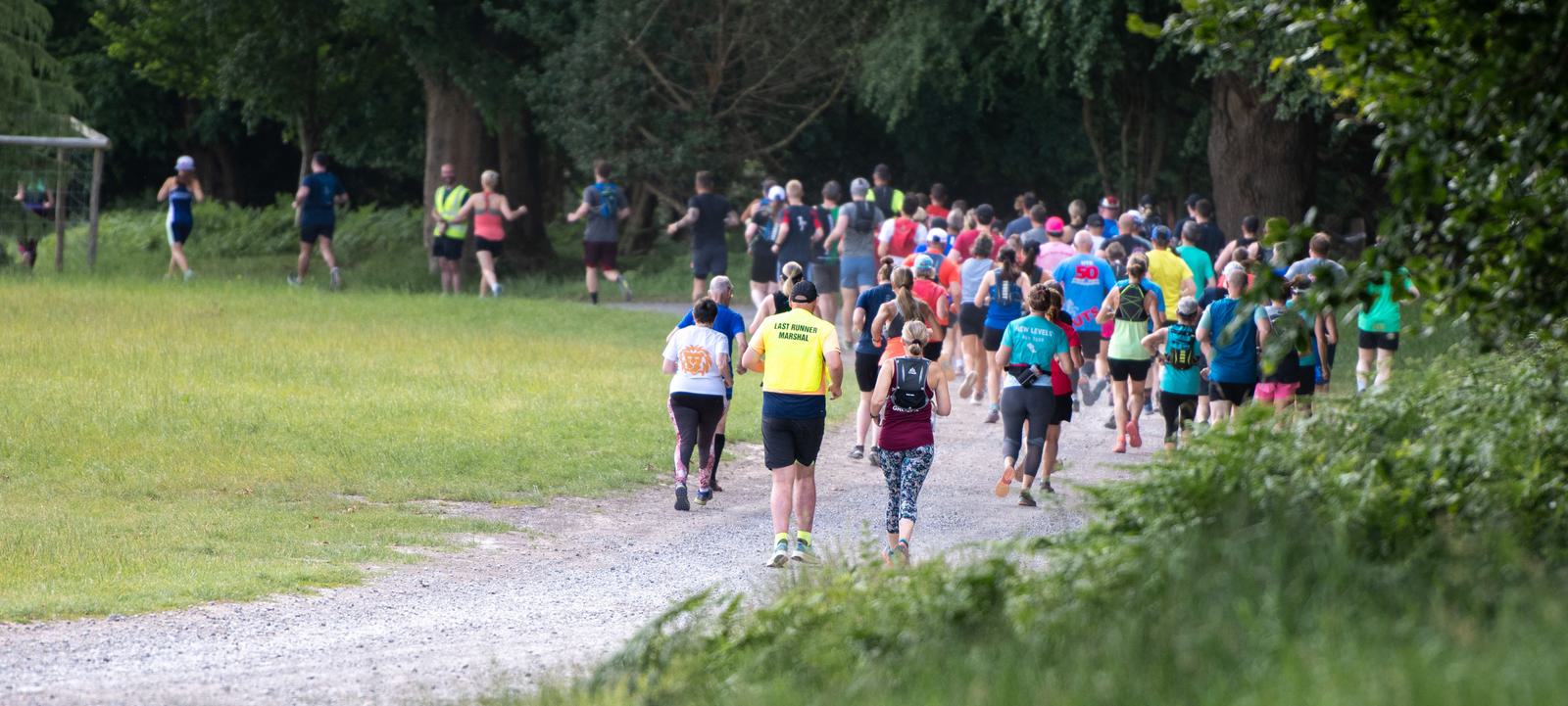 A large group of runners is participating in an outdoor race on a gravel path. They are surrounded by greenery and trees, with some runners wearing bright shirts and vests. The scene conveys a sense of community and activity in nature.