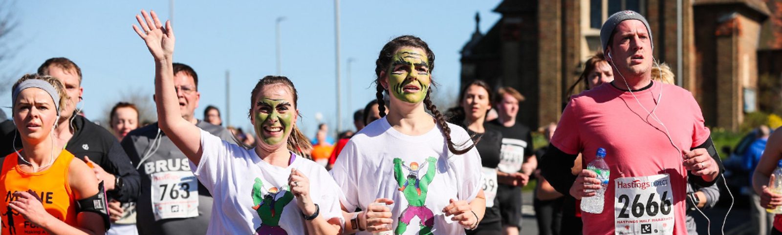A group of runners participate in a race. Two women in the center wear white shirts and have green face paint, while the man beside them wears a pink shirt and carries a water bottle. Other runners are visible in the background.