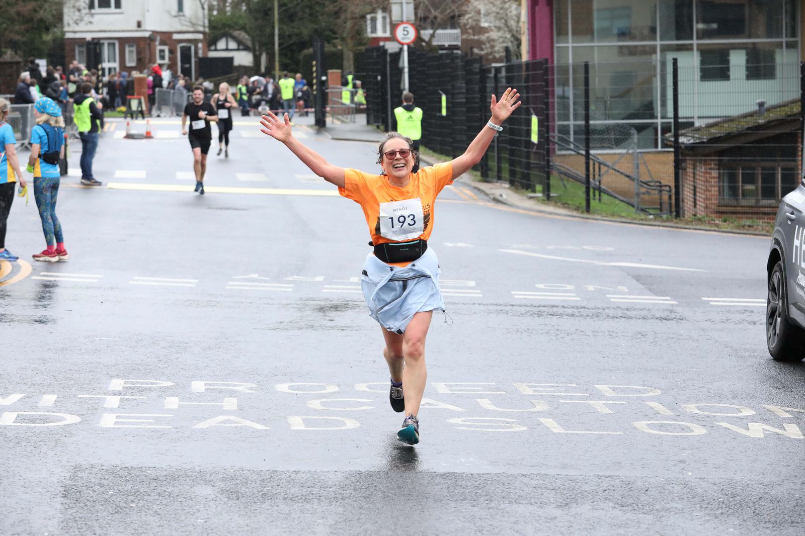 A person wearing an orange shirt and shorts is running with arms raised and smiling during a race on a city street. Other runners are visible in the background, as well as marshals and spectators. The street is wet, suggesting recent rain.