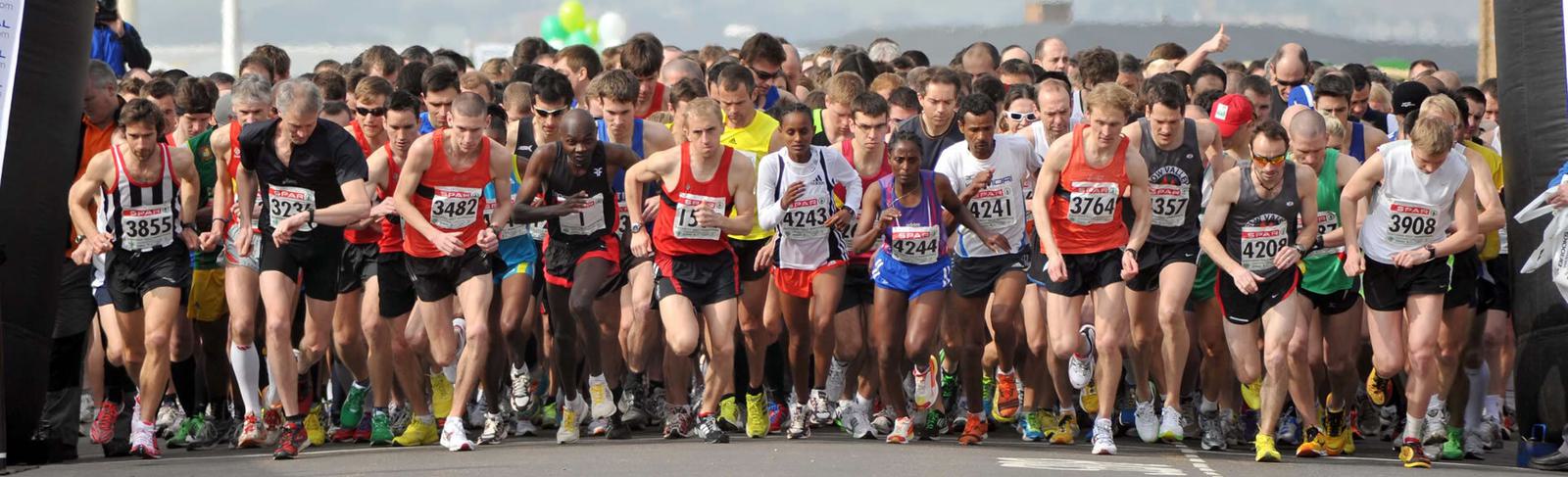 A large group of runners wearing various athletic gear and numbered bibs starts to run at the beginning of a race on a wide street. The background shows a cloudy sky and some distant landscape features.