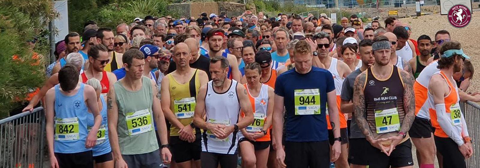 A large group of runners stands at a race starting line, each wearing numbered bibs and athletic gear. They are focused and preparing for a race. The background shows outdoor scenery with trees and fencing.