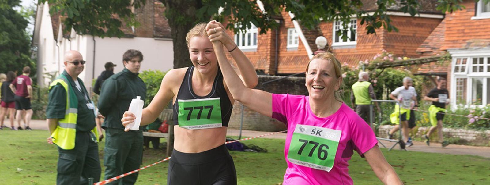 Two women, wearing race bibs 777 and 776, joyfully cross the finish line of a 5K run, holding hands. They appear enthusiastic and happy. Spectators and officials are visible in the background near a brick building.