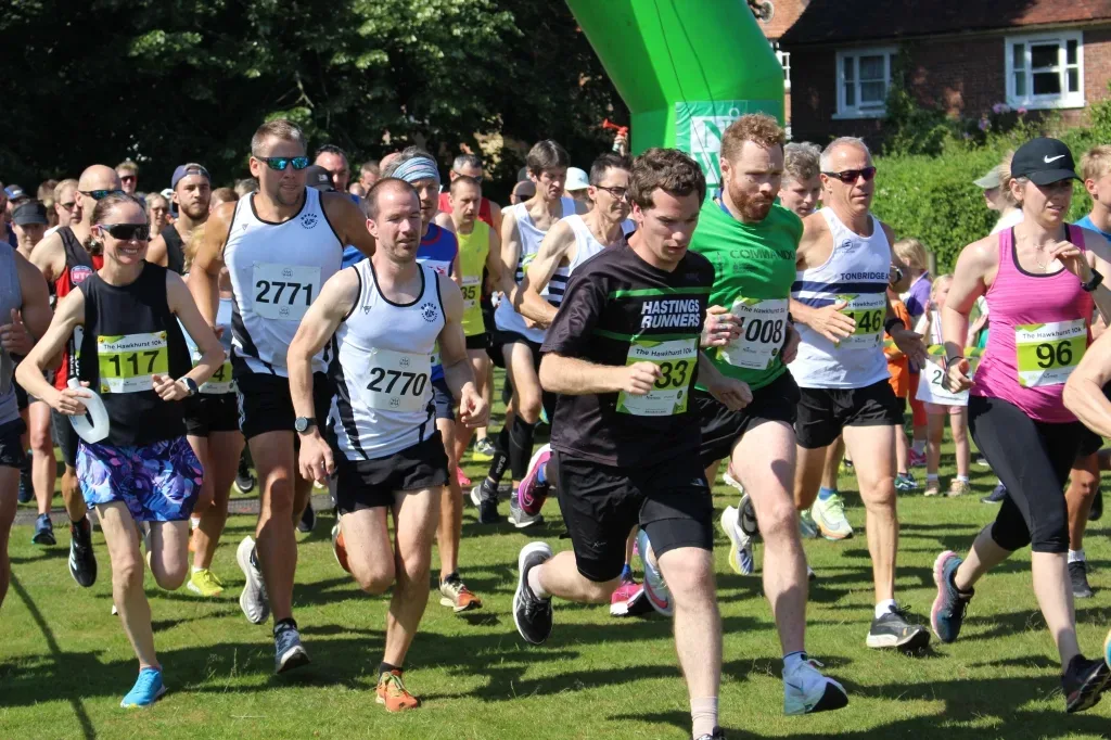 A group of runners, wearing race bibs and athletic gear, start a race on a sunny day. They are on a grassy field with trees and houses in the background. Some runners are mid-stride, while others are just beginning to run. The race environment appears energetic.