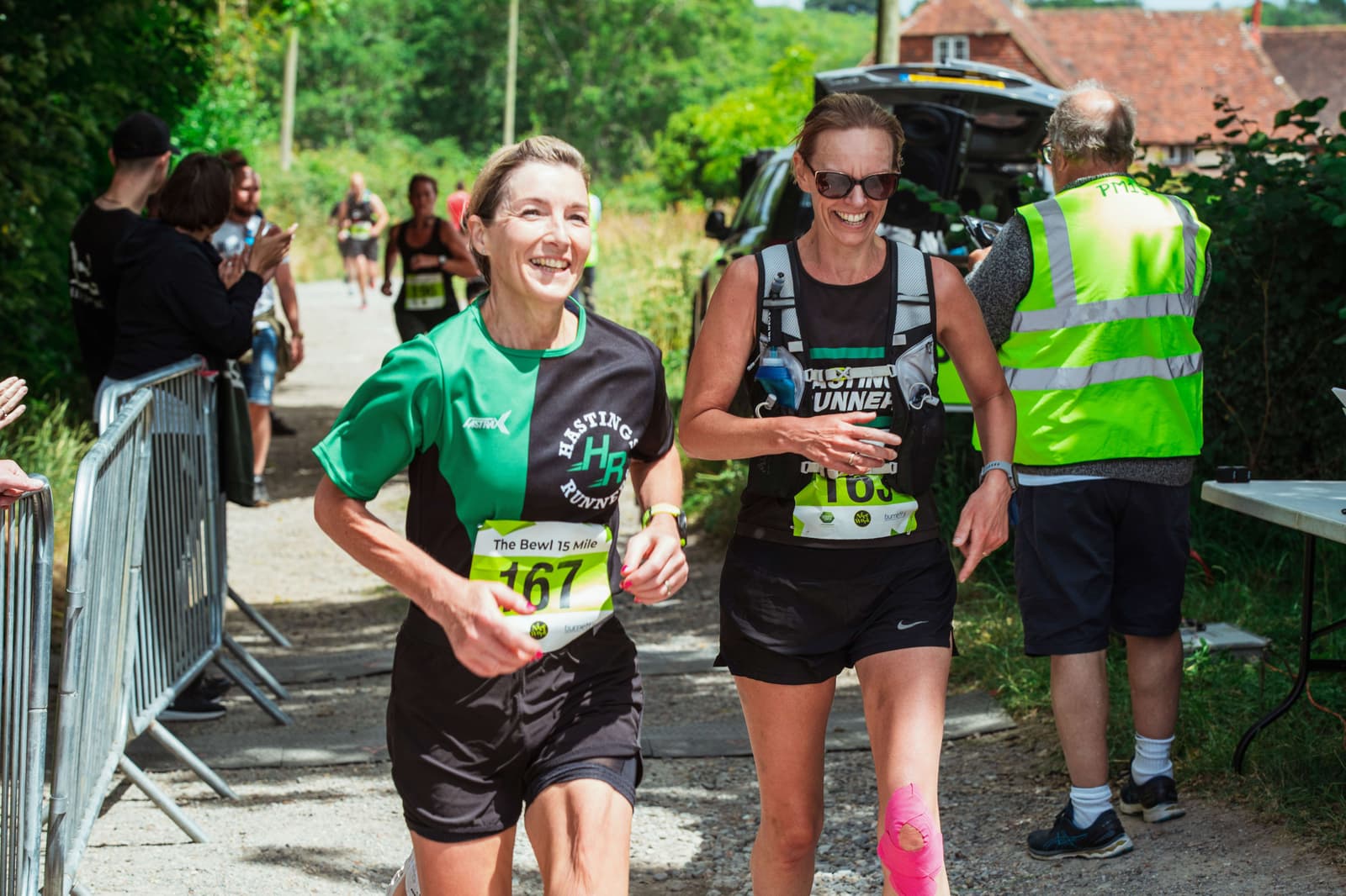 Two women, smiling and wearing running gear with race numbers, cross a race finish line outdoors. One wears a green top, the other black. People cheer in the background and a volunteer in a yellow vest stands nearby.