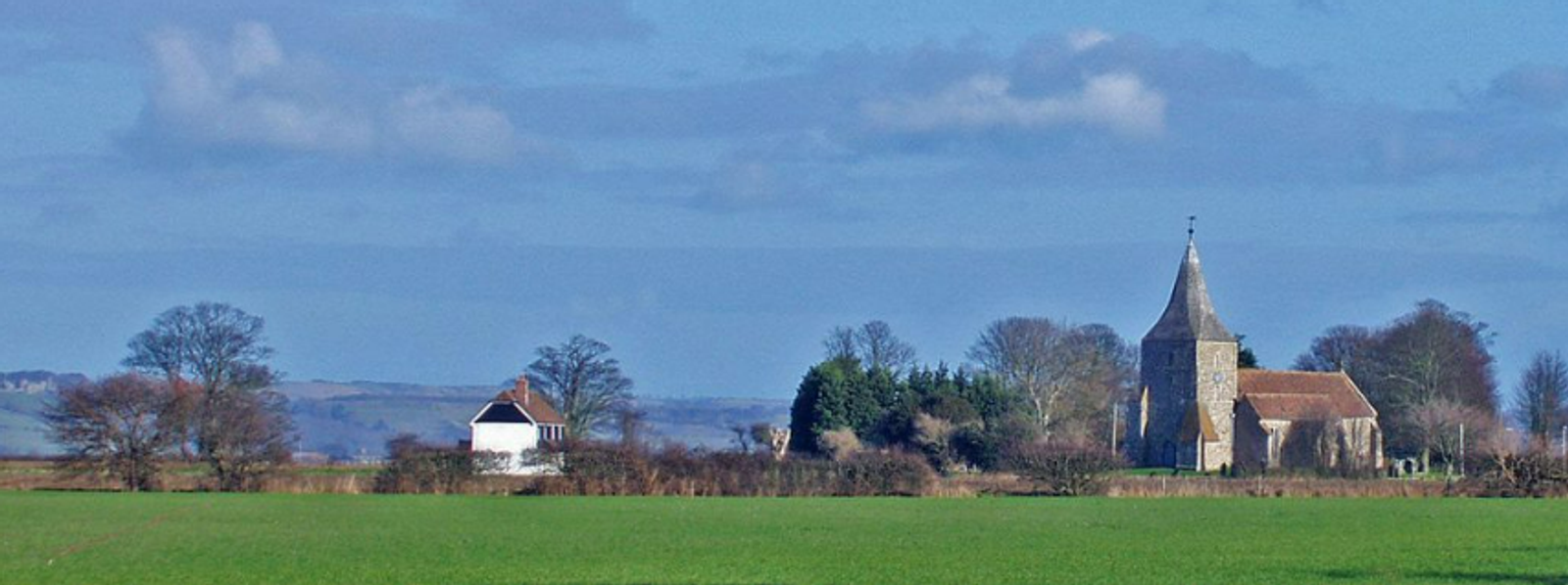 A tranquil rural landscape featuring a small stone church with a pointed roof surrounded by trees on the right, and a white house with a red roof on the left. Rolling hills and wide open skies complete the serene scene.