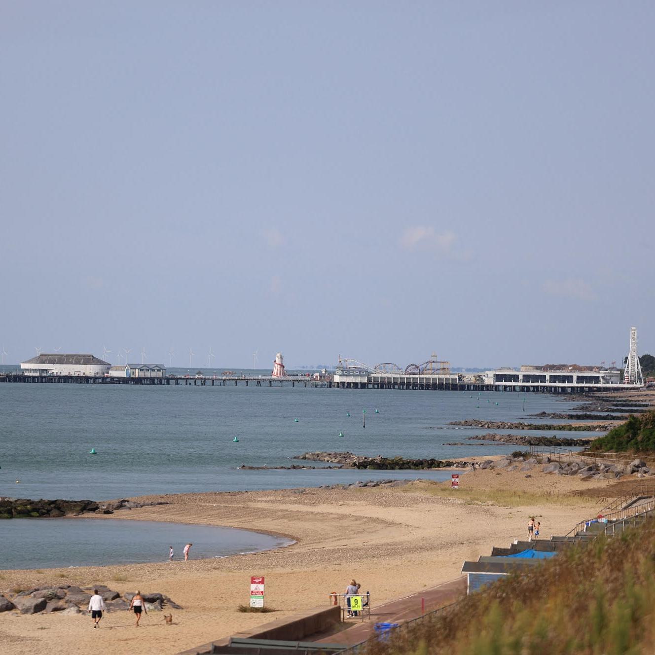 A beach scene with a few people walking along the sandy shore. The calm sea stretches out to the horizon, and a long pier with buildings stands in the distance under a clear blue sky.