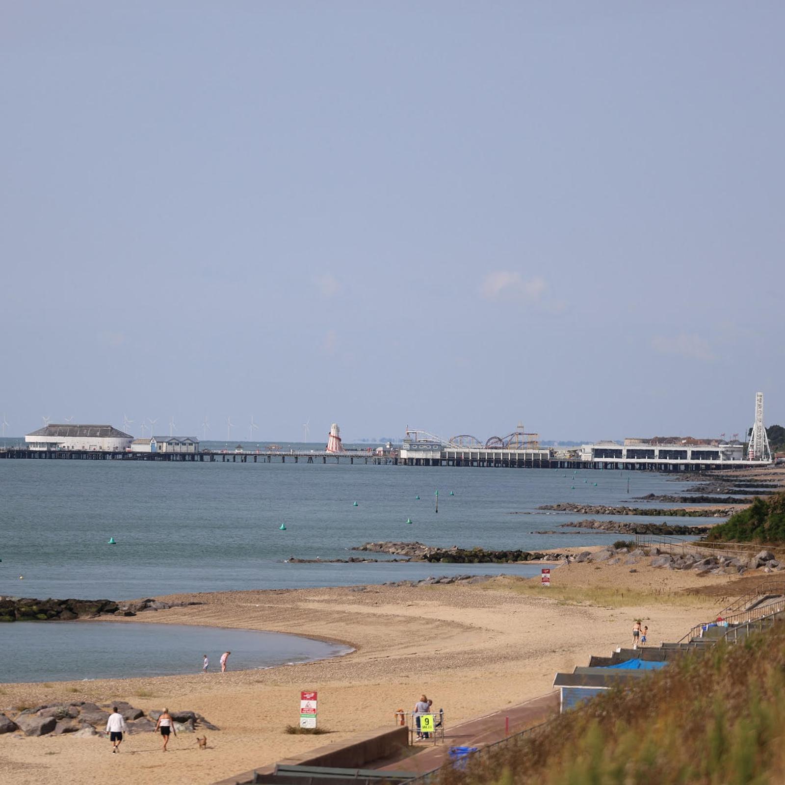 A beach scene with a few people walking along the sandy shore. The calm sea stretches out to the horizon, and a long pier with buildings stands in the distance under a clear blue sky.
