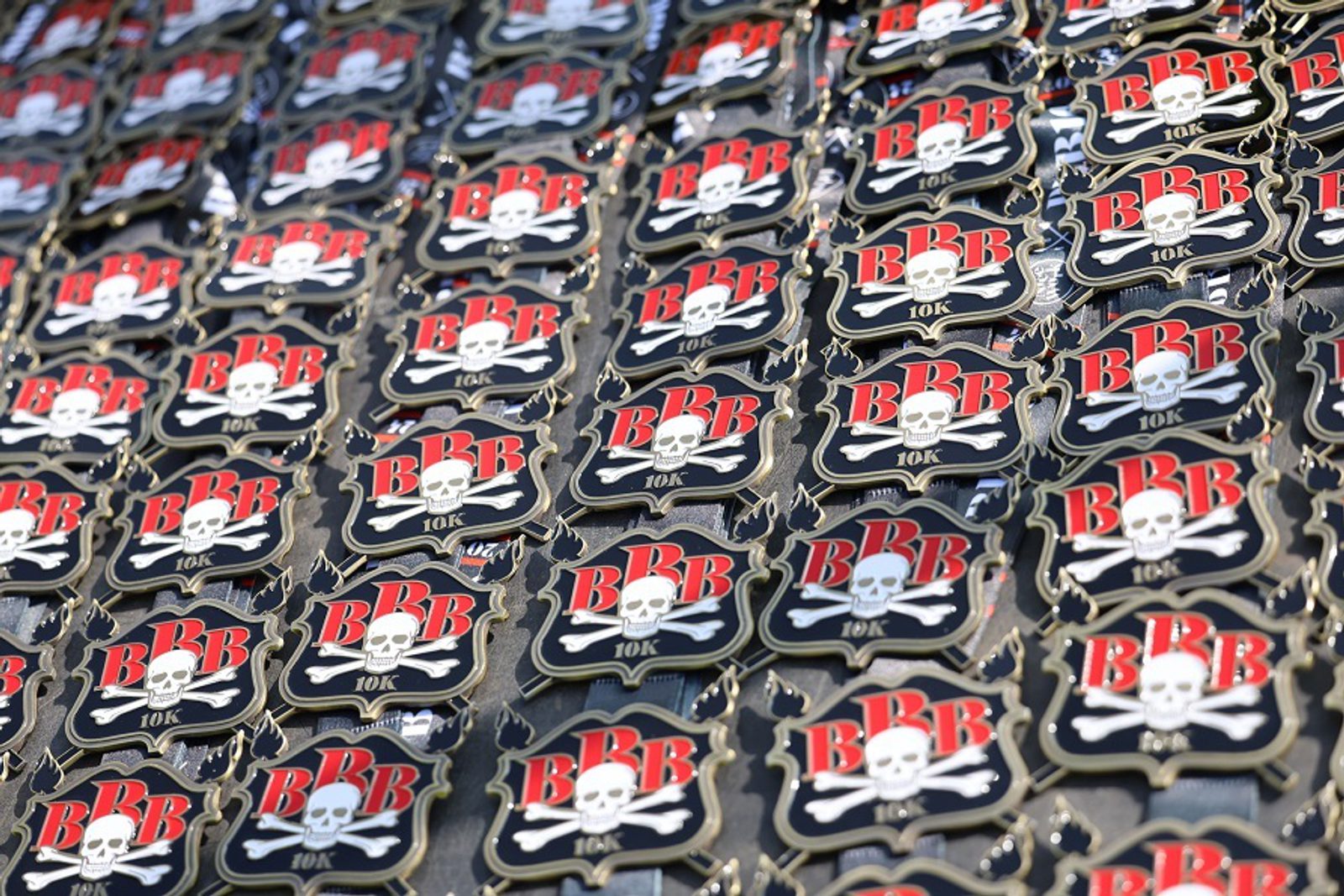 Rows of black and gold medals featuring a skull and crossbones, the letters “BBB”, and “10K” text, arranged closely together in a repeating pattern.
