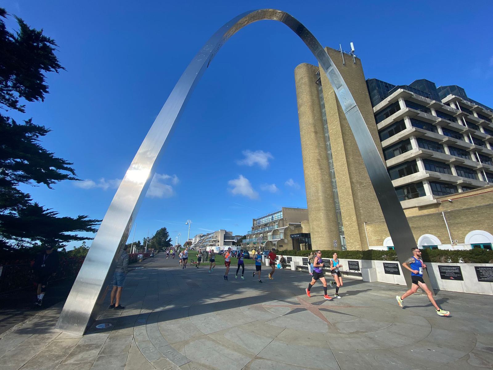Joggers run under a large, silver archway on a bright day. The scene includes modern buildings, a clear blue sky, and a few pedestrians enjoying the outdoor environment.