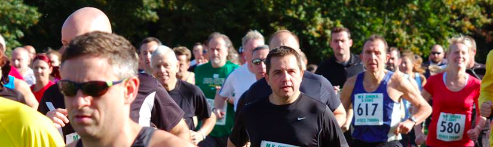 A group of runners participating in a race, wear race bibs with numbers. They are running on a road bordered by trees. The runners are of various ages and displayed in a tightly packed formation, with some wearing sunglasses and others in colorful athletic gear.