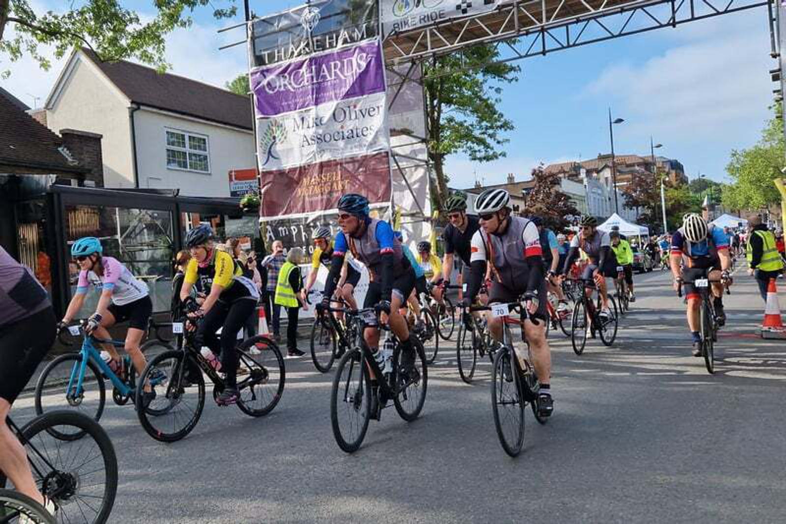 Cyclists participating in a road race, wearing helmets and gear, pass under a banner on a sunny day. Spectators and race officials are visible in the background along a street lined with trees and buildings.