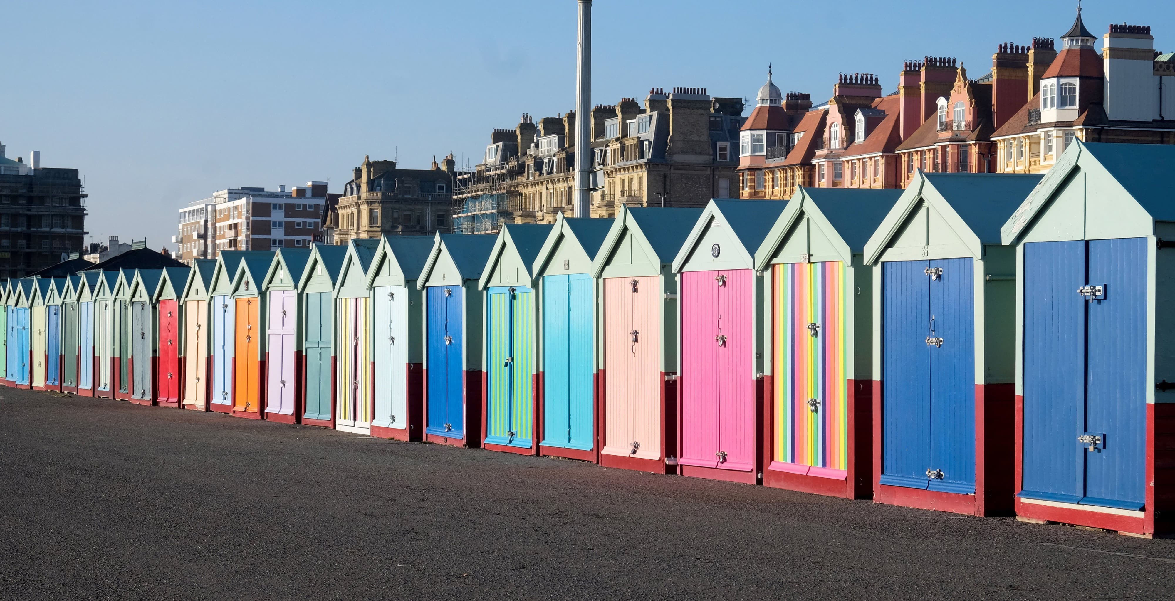 A row of colorful beach huts with pastel blue, green, pink, and yellow doors stands in front of historic multi-story buildings under a clear blue sky.