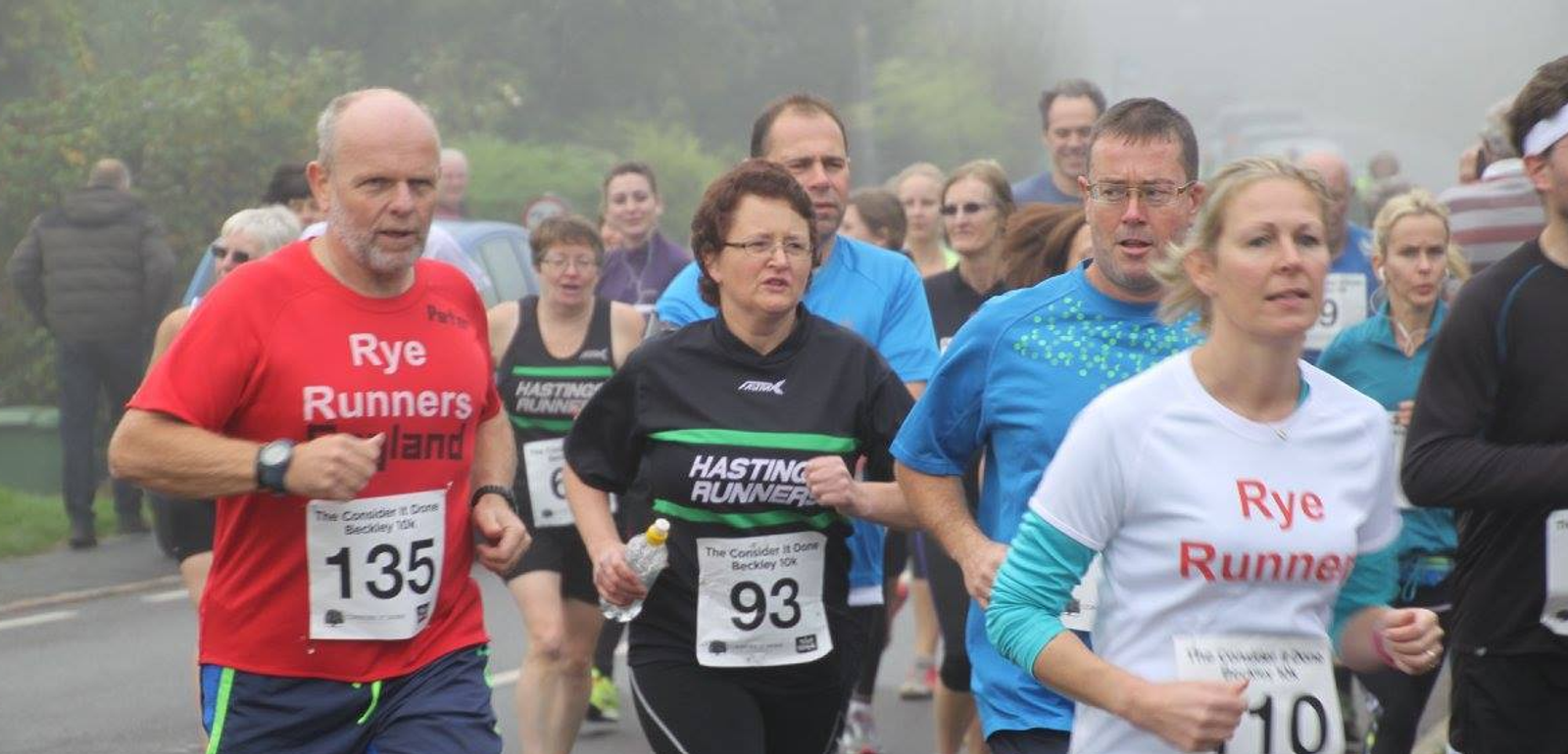 A group of runners participating in a road race, all wearing numbered bibs. The runners are of varying ages and are dressed in colorful athletic clothes. The environment appears to be misty, with greenery in the background. Some runners wear shirts with club names.