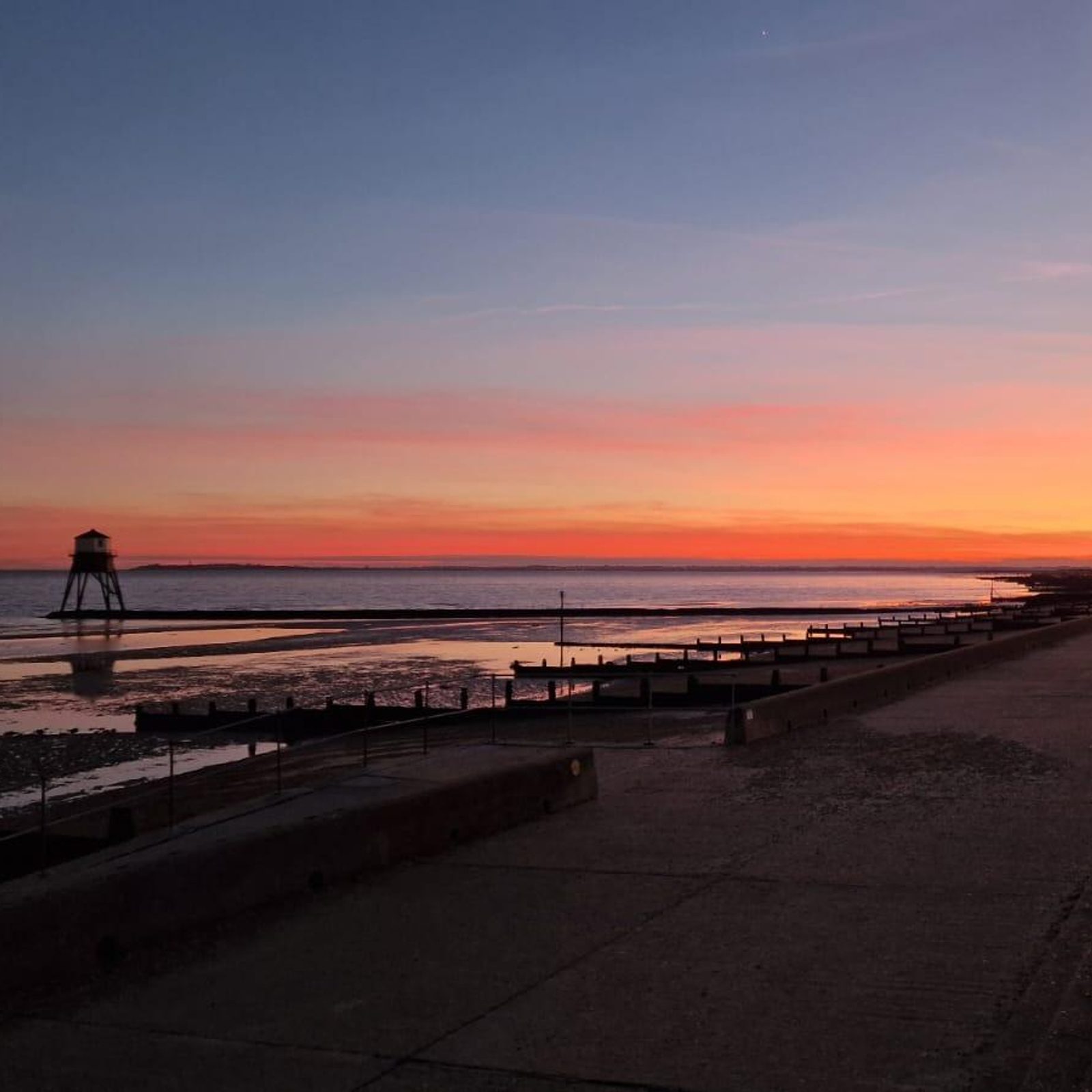A tranquil beach at sunset with a vibrant orange and pink sky. A wooden lifeguard tower stands on the left near the shore, and a concrete promenade runs along the right side of the image.