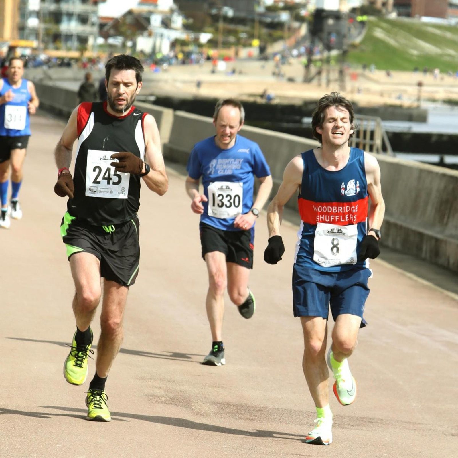 Three male runners wearing race bibs compete in a road race near a seafront promenade, with other runners and the beach visible in the background. The weather appears sunny and clear.