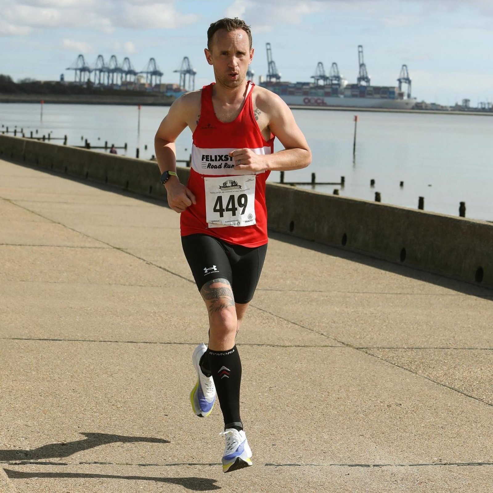 A man wearing a red tank top and black shorts is running in a race along a paved waterfront path. He has a race bib numbered 449 and there are cranes visible in the background across the water.