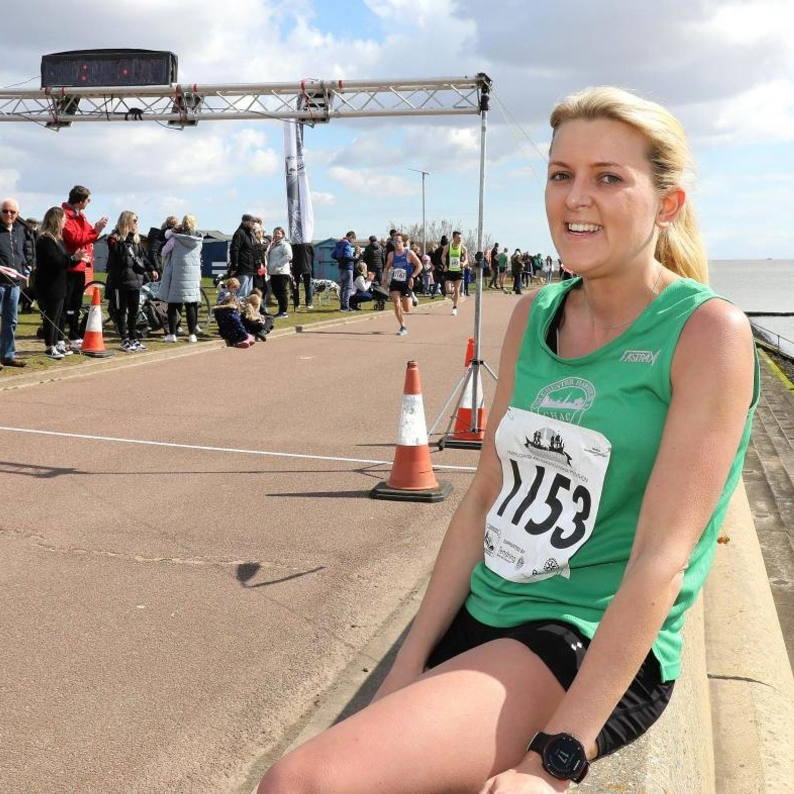 A smiling woman in a green tank top with race number 1153 sits on a curb after a road race. Spectators and other runners are in the background near a finish line by the water.