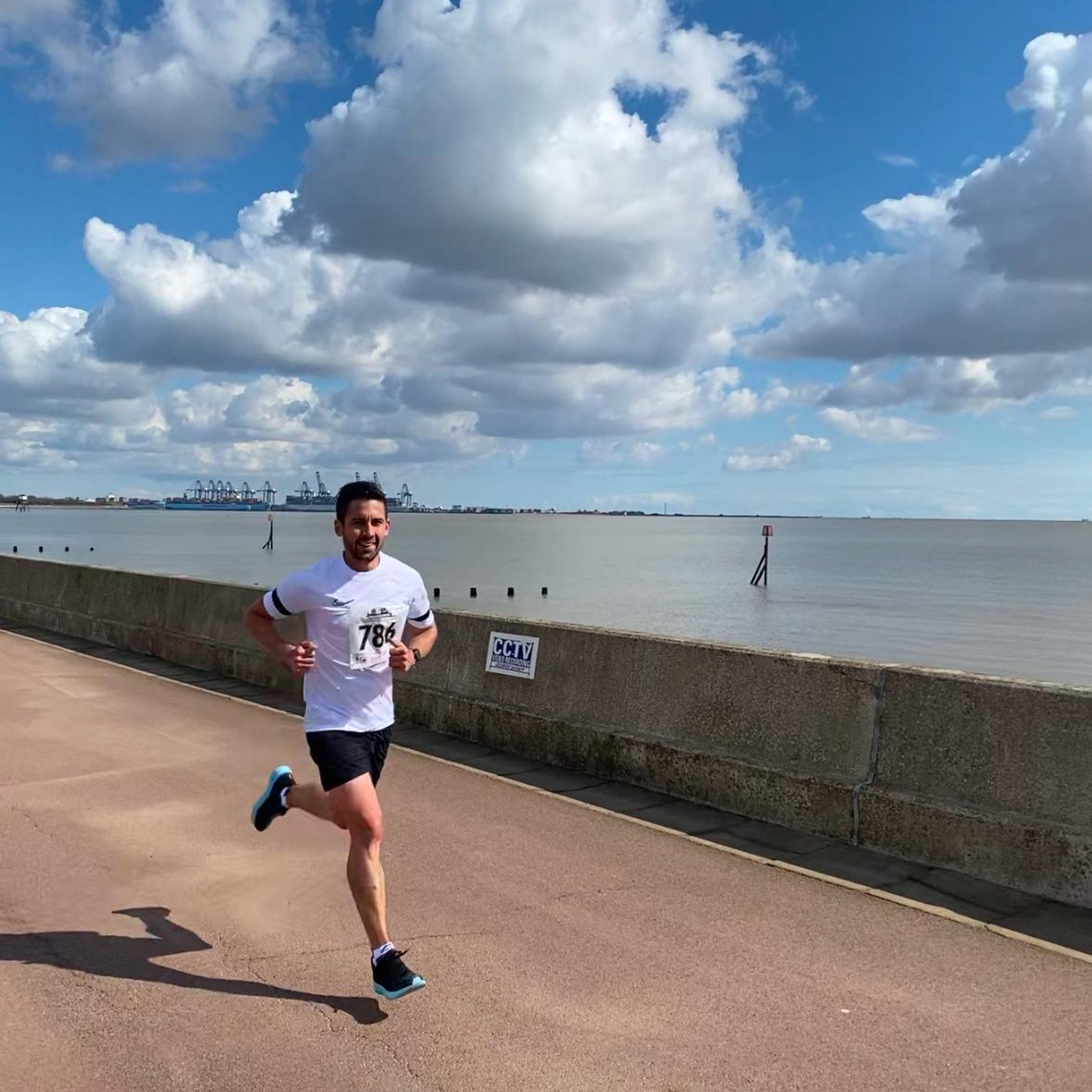 A man in a white t-shirt and black shorts is running along a seaside promenade under a blue sky with scattered clouds. The calm sea and distant industrial structures are visible in the background.