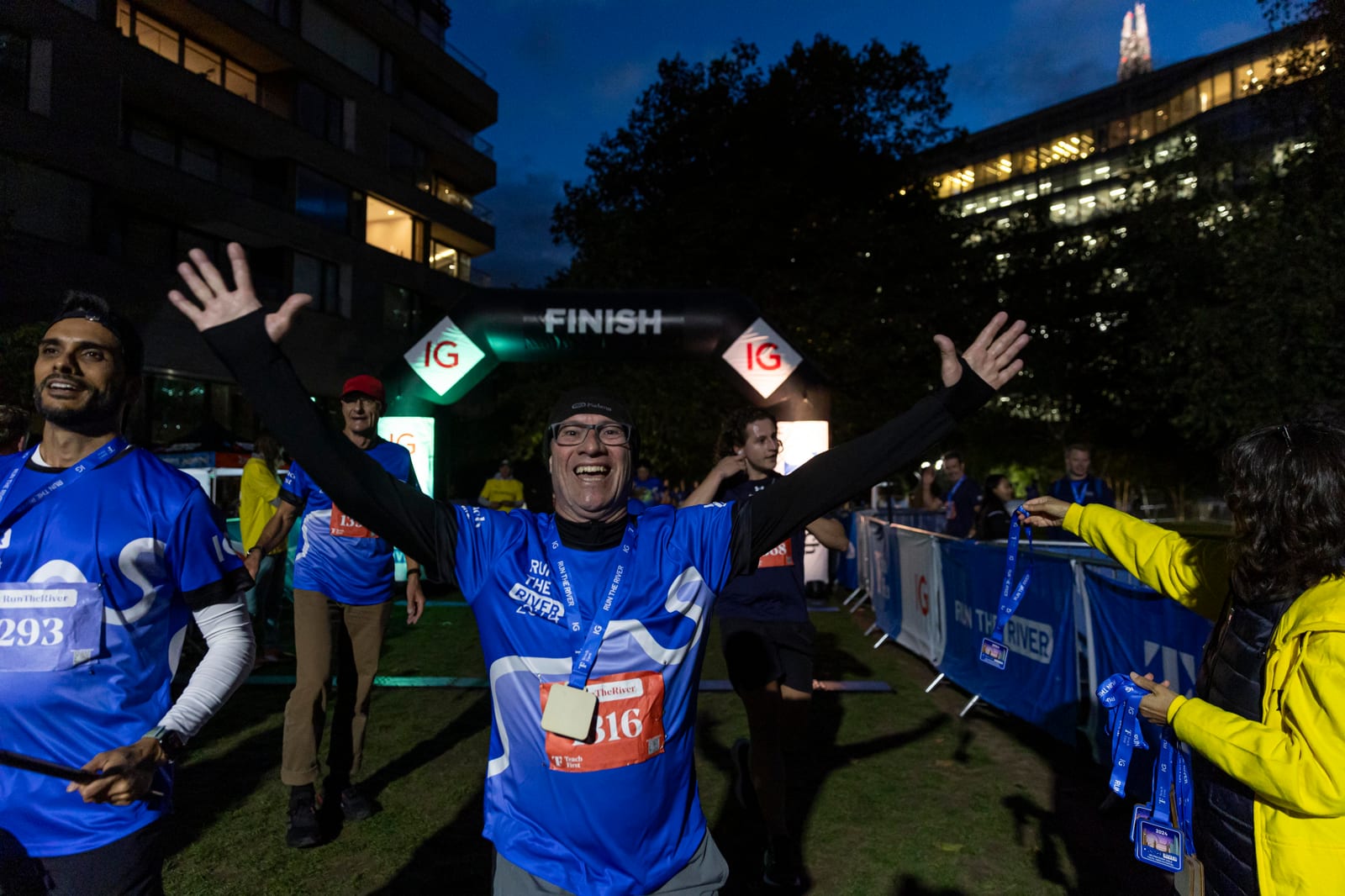A smiling runner in a blue shirt celebrates with arms raised at a nighttime race finish line, while others stand nearby and a medal is being handed out. The FINISH banner is visible in the background.