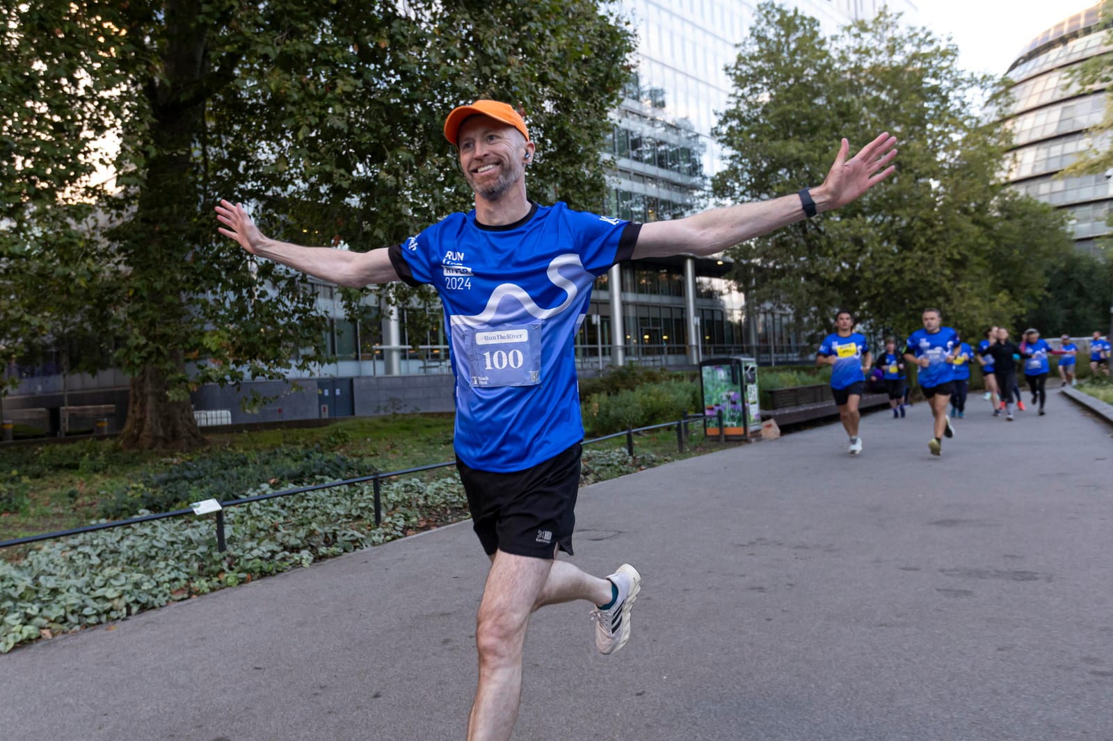 A smiling runner in a blue shirt and orange cap spreads his arms wide while running on a city park path, with other runners and tall buildings in the background.