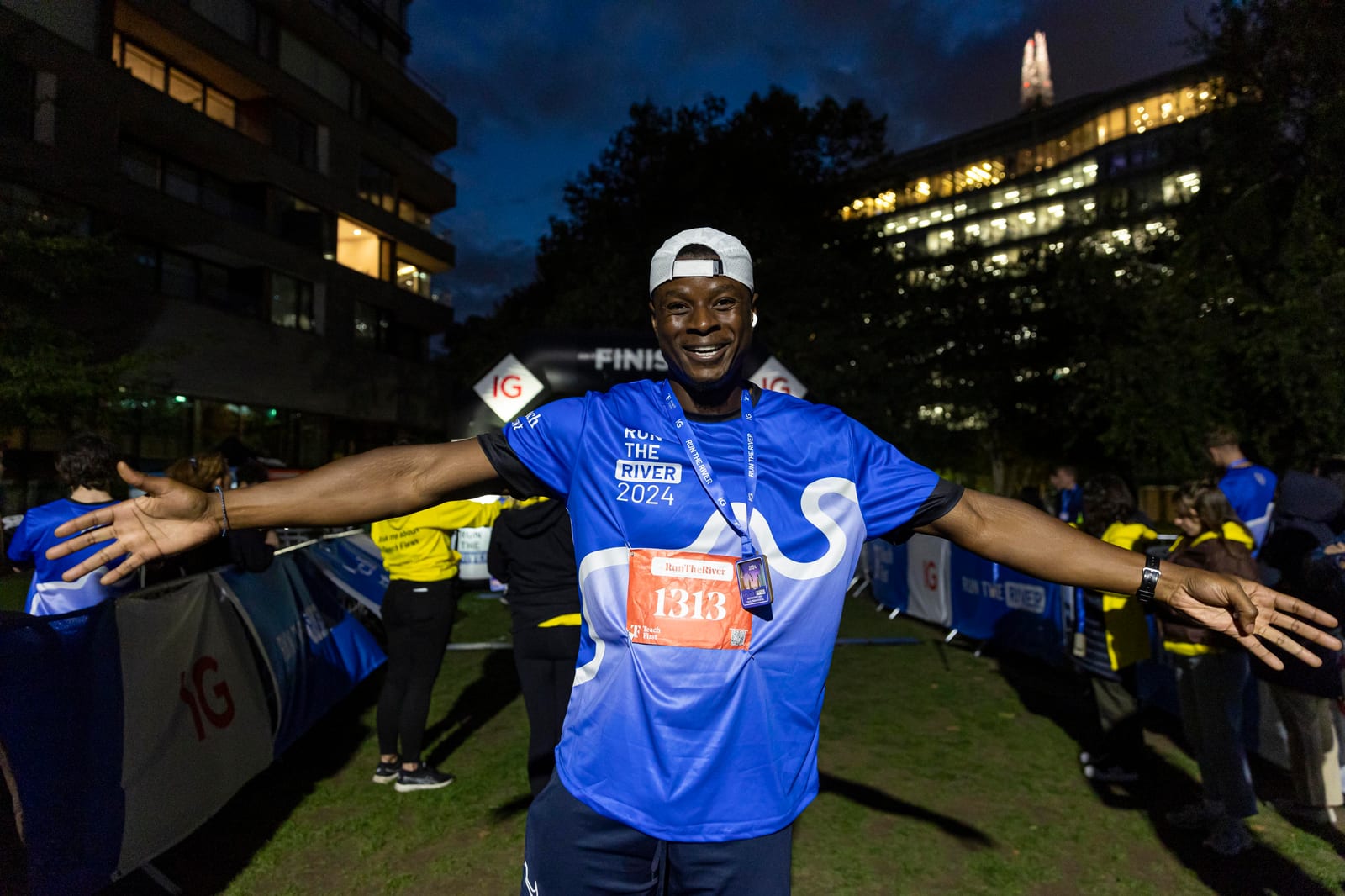 A smiling runner wearing a blue "Run the River 2024" shirt and race number 1313 spreads his arms wide at the finish line of a nighttime race event, with other participants and city buildings in the background.