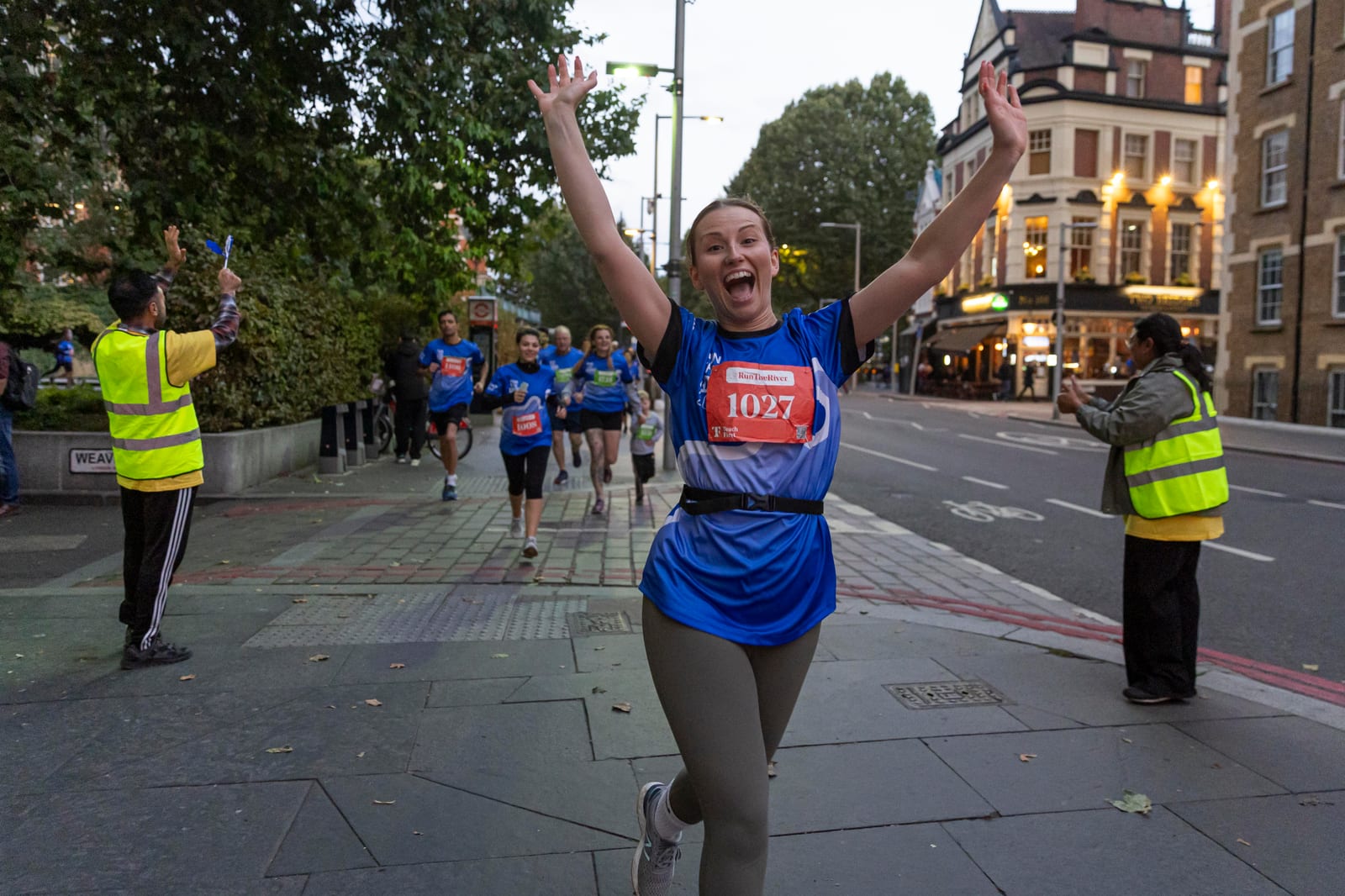 A woman in a blue shirt with race number 1027 raises her arms and smiles while running on a city street during a race, with other runners and volunteers in yellow vests visible in the background.