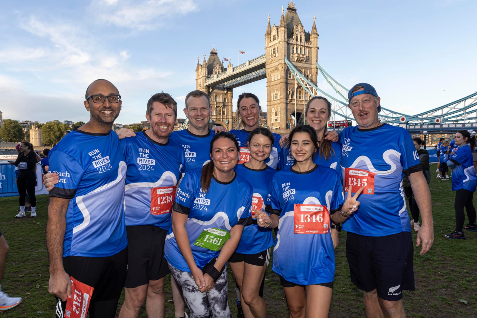 A group of smiling runners in blue "Run the River 2024" shirts pose together in front of Tower Bridge, holding their race bibs, on a grassy area during daylight.