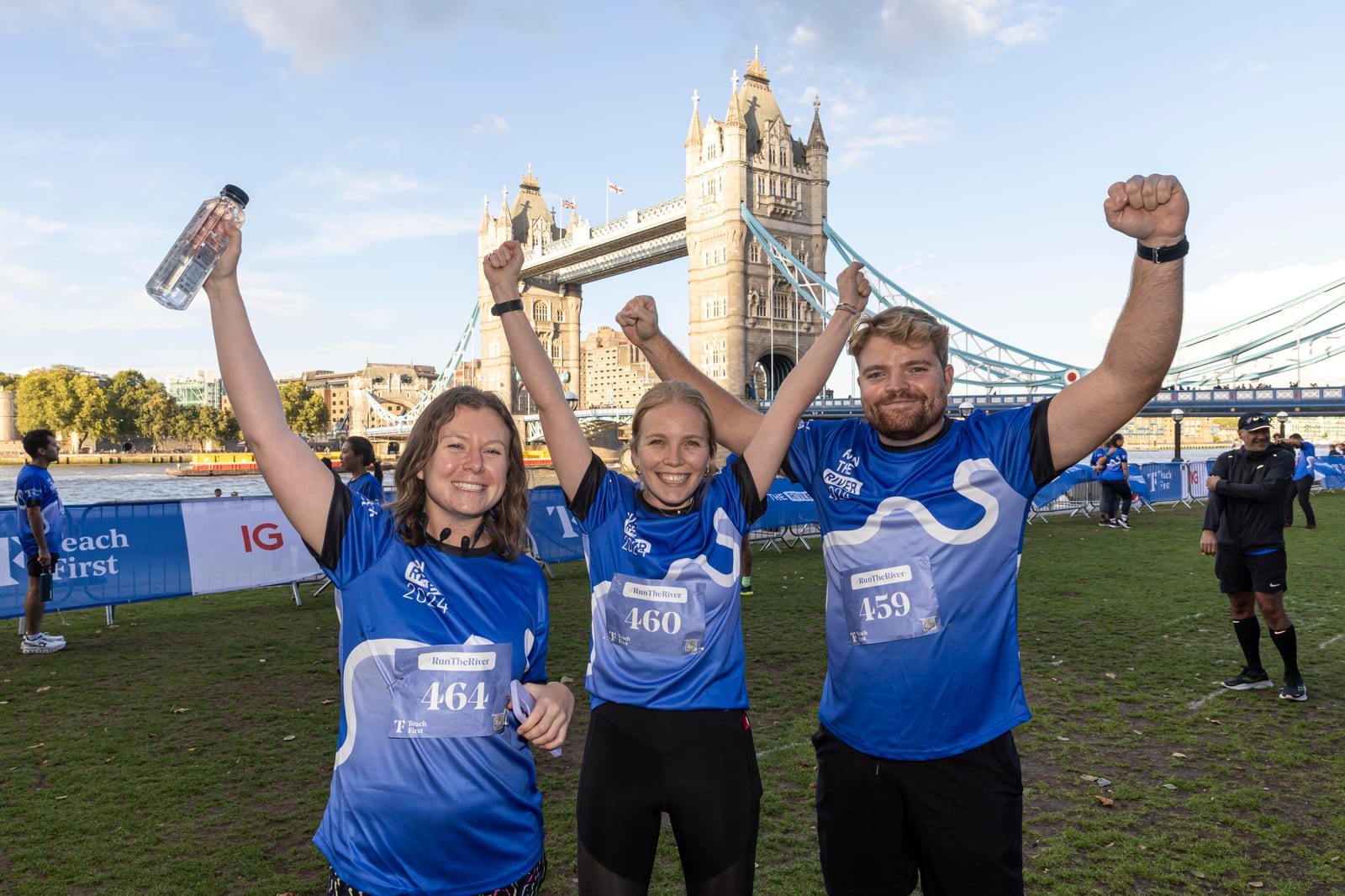 Three smiling runners in blue shirts raise their arms in celebration after a race, standing on grass with Tower Bridge in the background on a sunny day. They wear race bibs and medals, and one holds a water bottle.